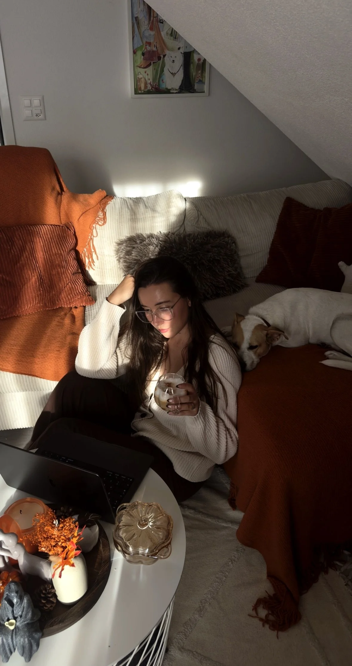 A woman with glasses and long dark hair relaxes on a couch in a cozy living room, holding a glass of wine. A dog is lying beside her on the couch. The room is decorated with orange and brown pillows, and a table with fall-themed decorations including candles, a pumpkin, and flowers.