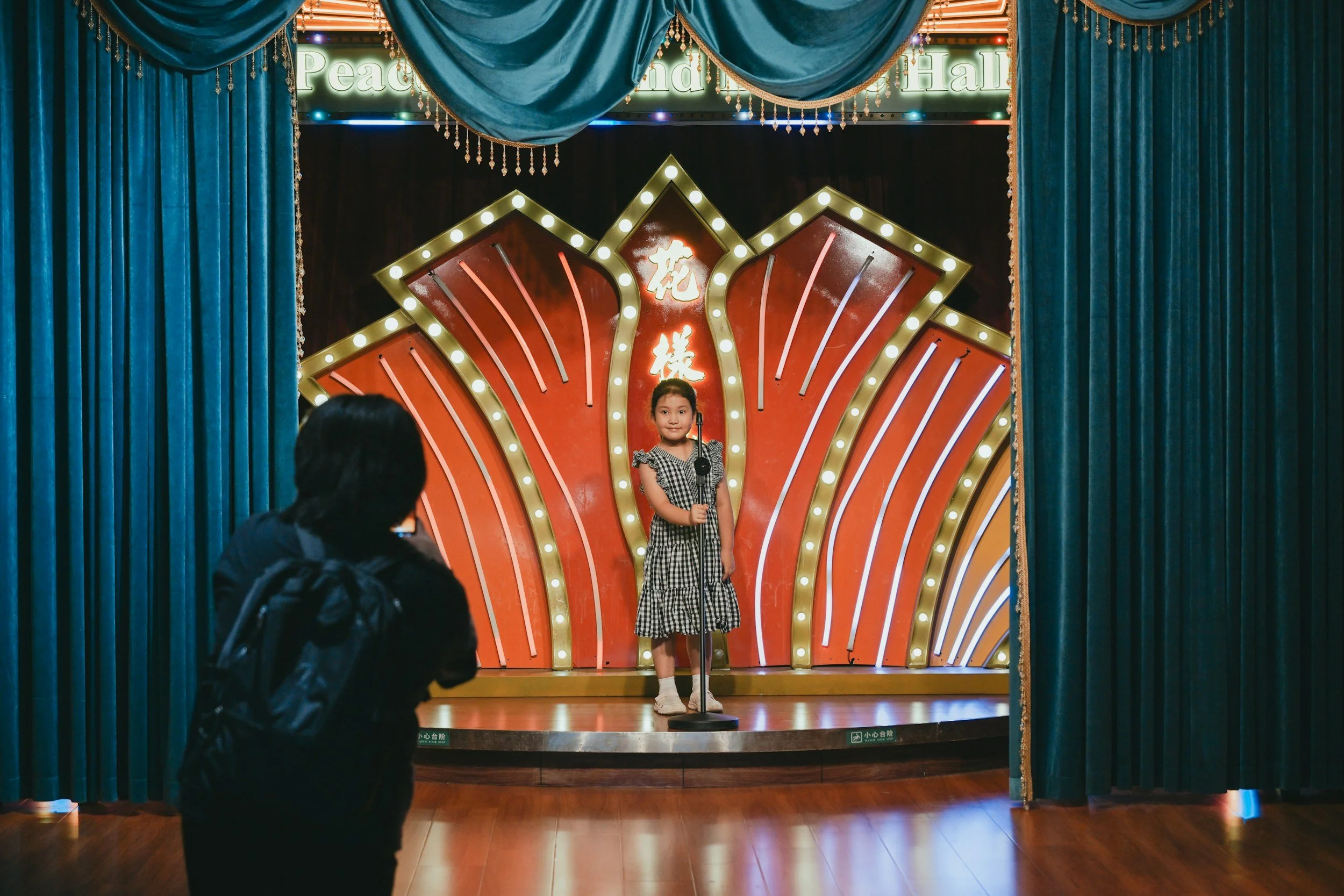 A young girl stands on a stage with a microphone, dressed in a black and white checkered dress, during a performance. The stage is decorated with a large, illuminated backdrop that has red and gold elements with Chinese characters, surrounded by blue curtains. Two adults are visible in the foreground watching the girl.