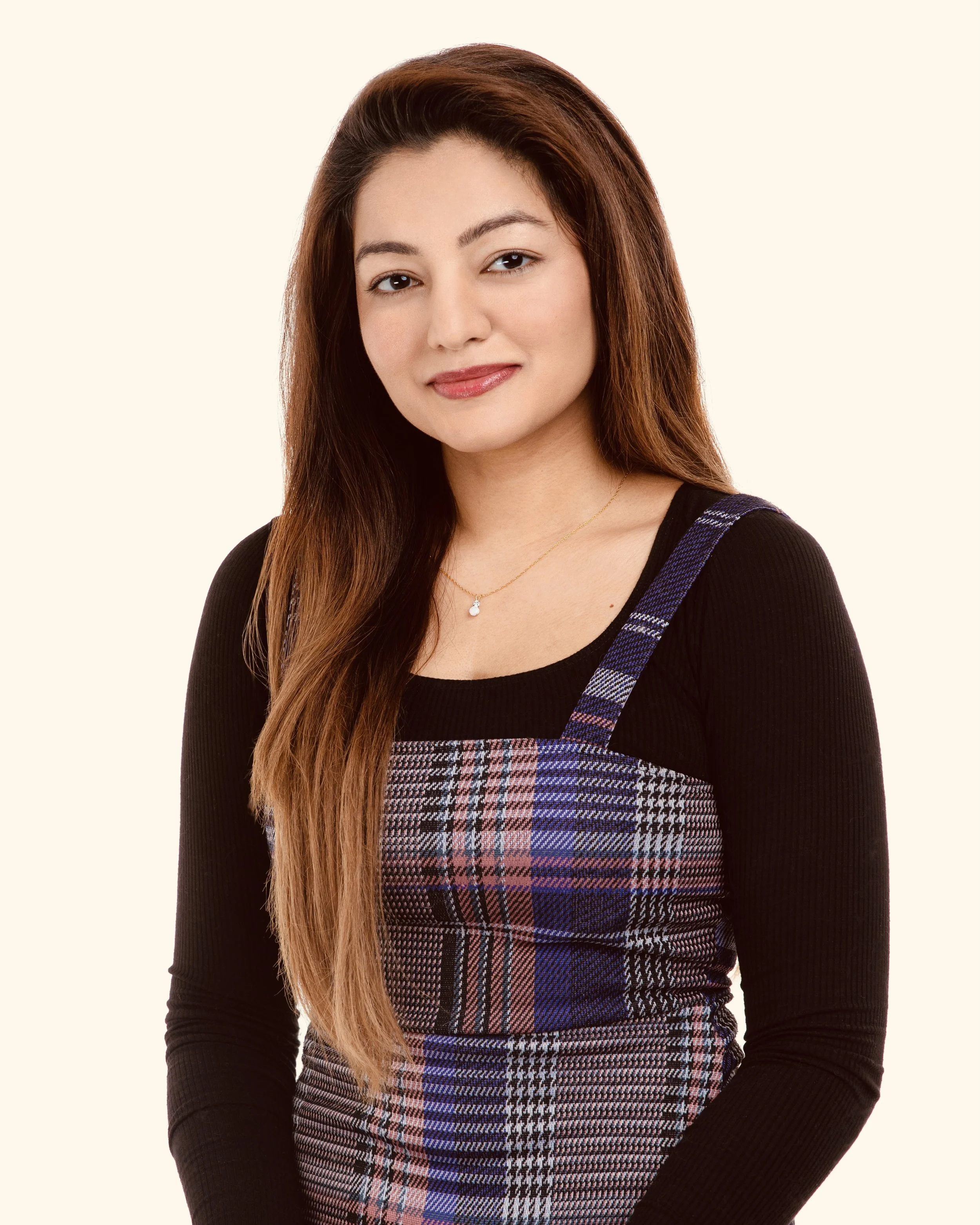 A young woman with long brown hair, wearing a black top with a plaid pinafore dress, and a delicate necklace, standing against a plain light background.
