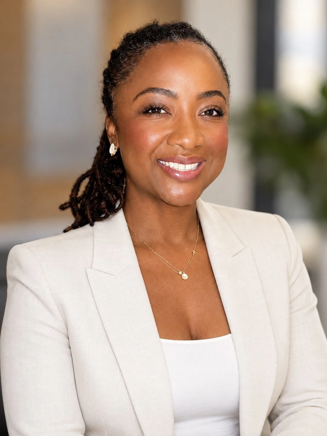 Professional woman smiling, wearing a white blazer and jewelry, in an indoor office setting.