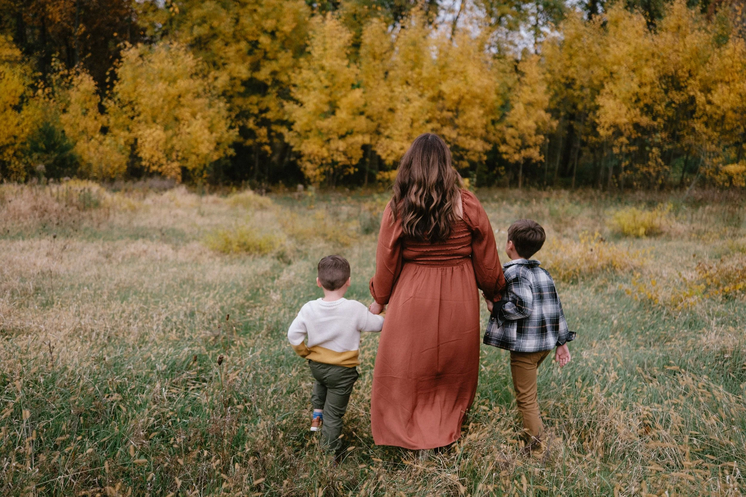 A woman walking with two children in a field during autumn, with trees in fall colors in the background.