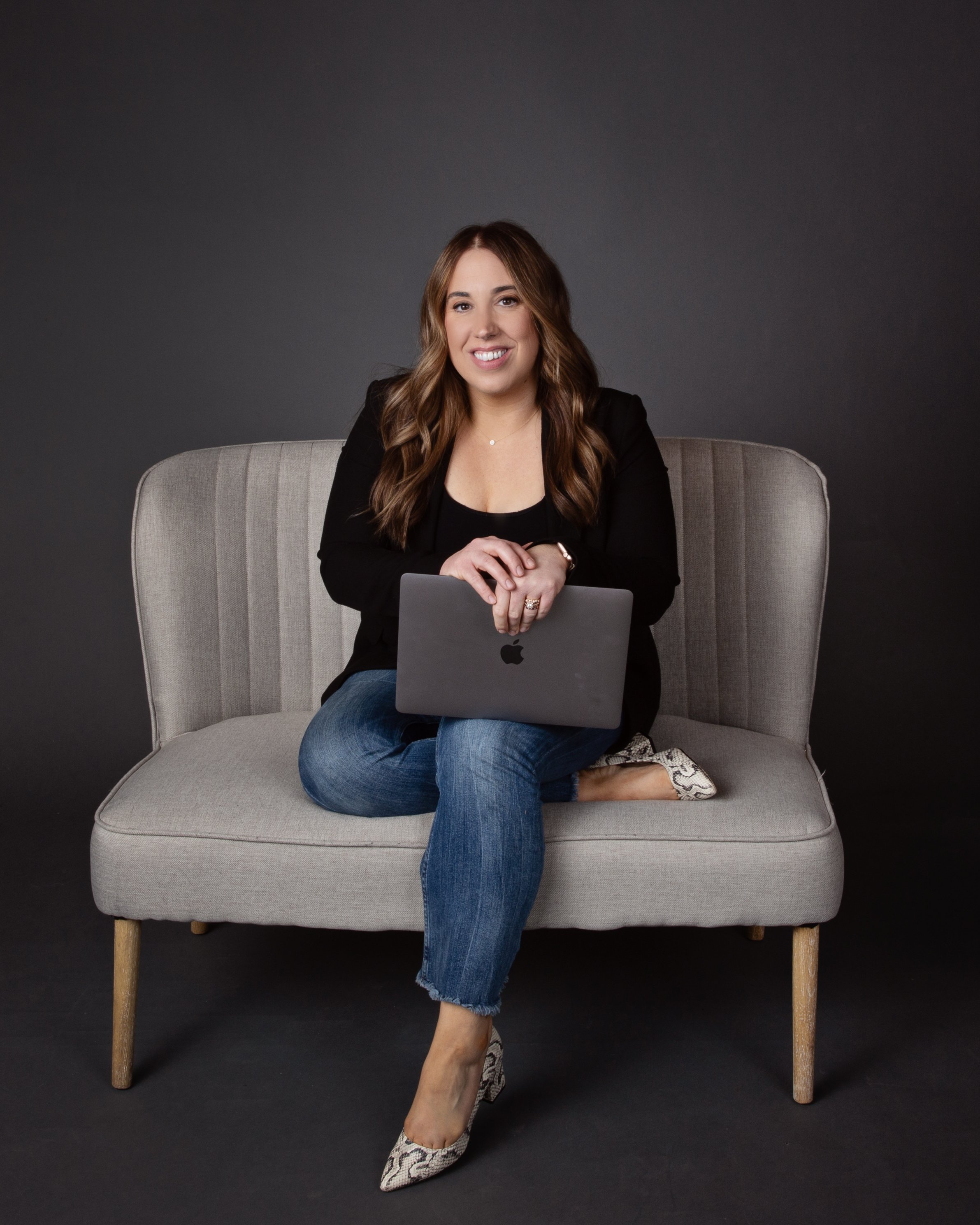 A woman with brown hair, wearing a black blazer, blue jeans, and snakeskin high heels, sitting on a beige sofa holding a closed gray MacBook.