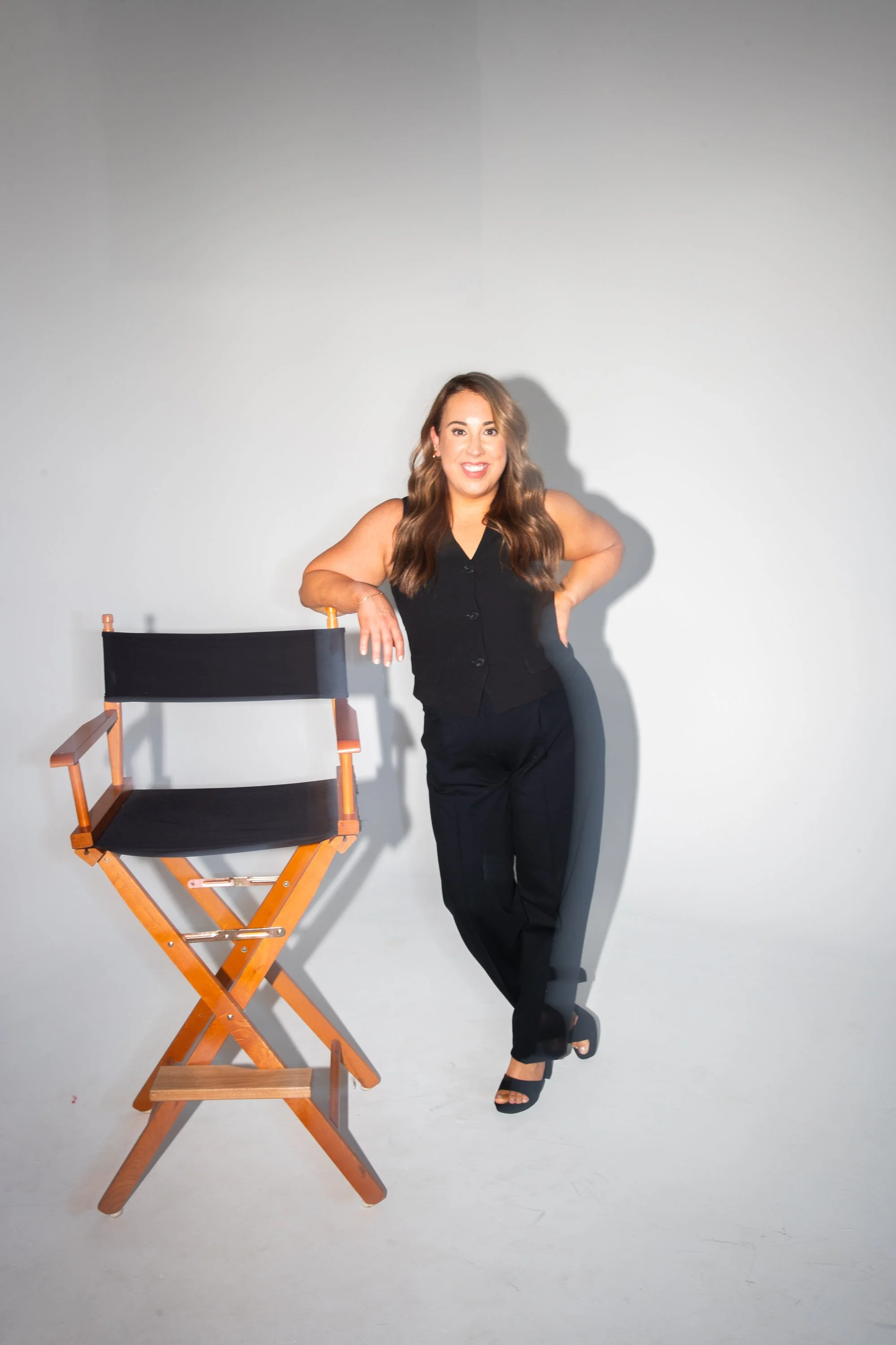 A woman with long brown hair, wearing a black sleeveless top and pants, smiling and standing next to a black director's chair with wooden armrests against a plain white background.