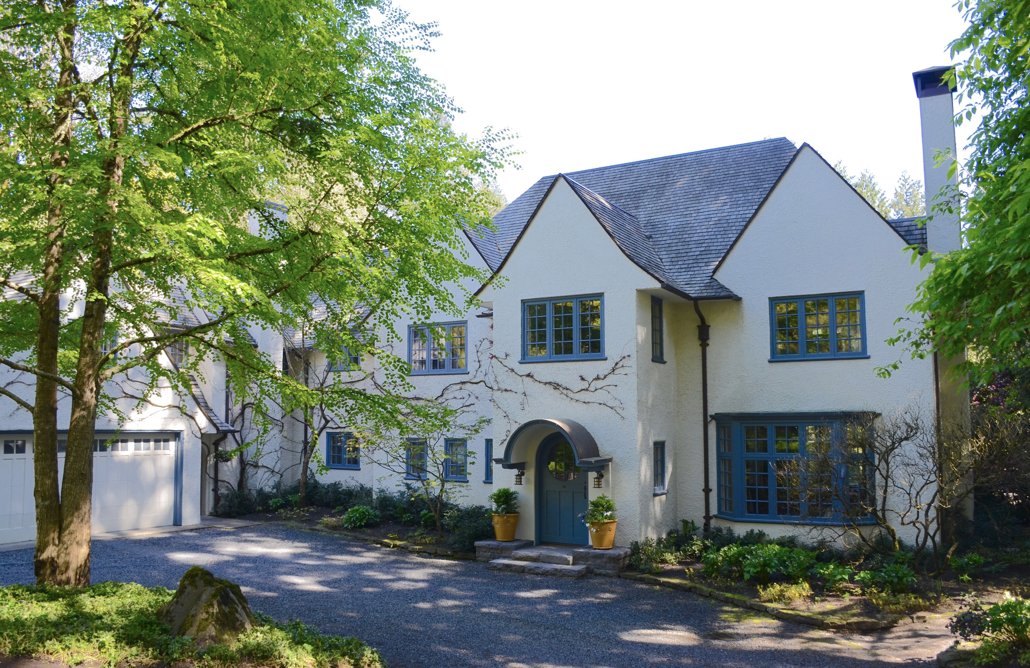 The Maurice Crumpacker House in Dunthorpe, Portland, a white stucco Wade Pipes house with steep gables, blue casement windows, a round-headed blue front door under an arched hood, a tall chimney and a gravel drive under trees.