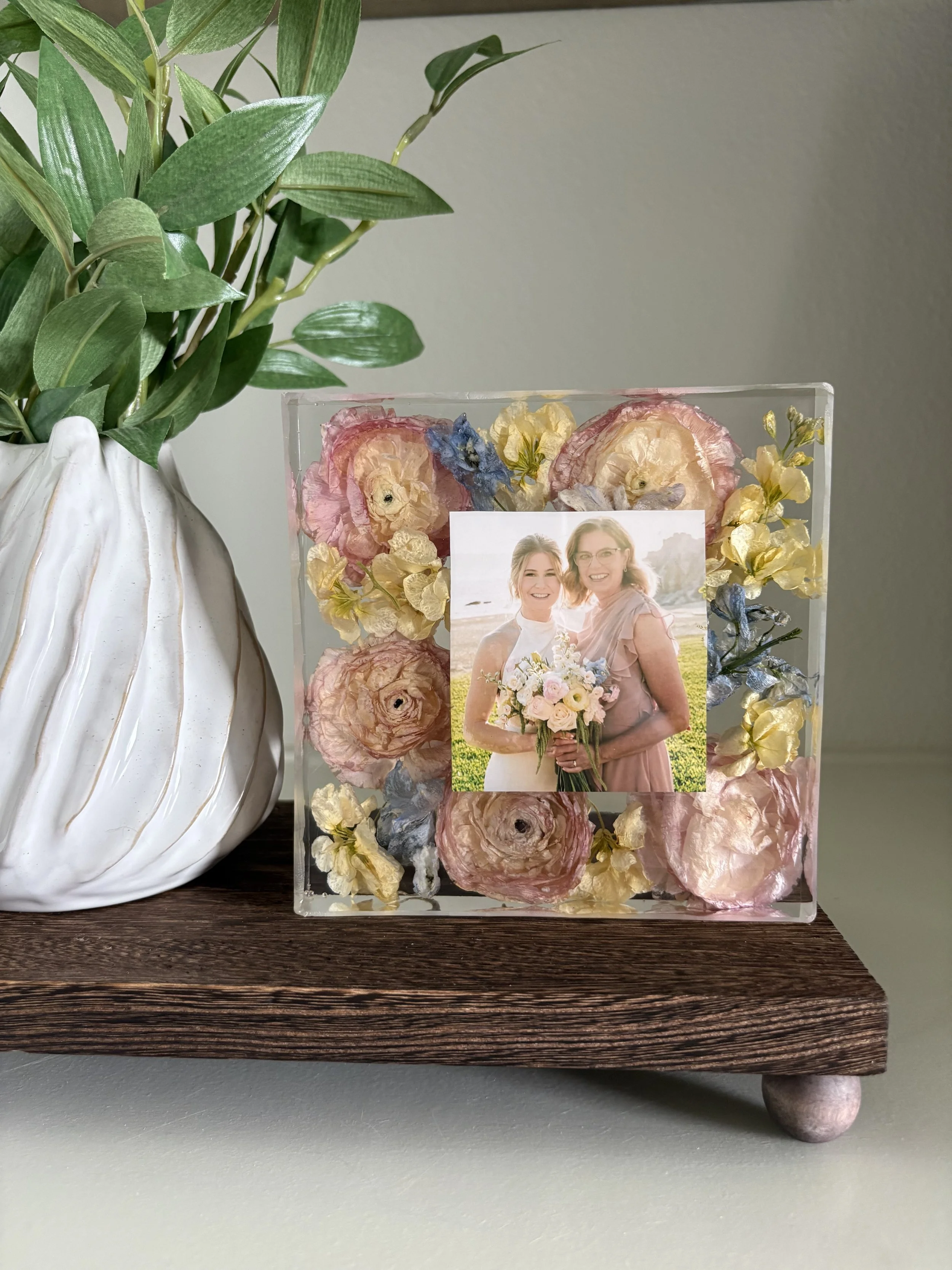 A vase with green leaves and a floral photo holder with pressed flowers, a small photograph of two women, and dried flowers, on a wooden surface.