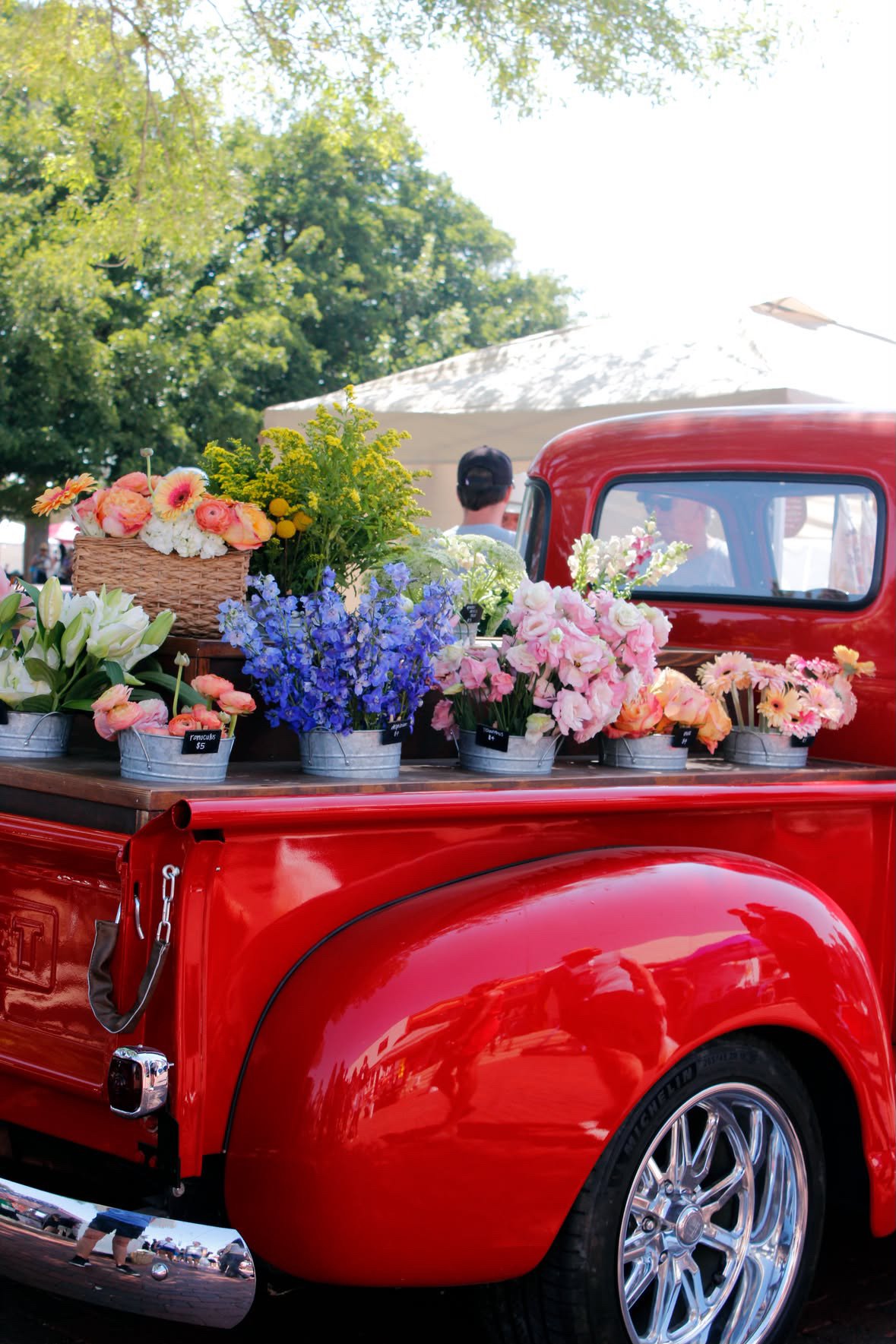 A vintage red pickup truck with a flatbed filled with various colorful flowers, including pink, white, purple, and yellow blooms, parked outdoors under a canopy of green trees during daytime.