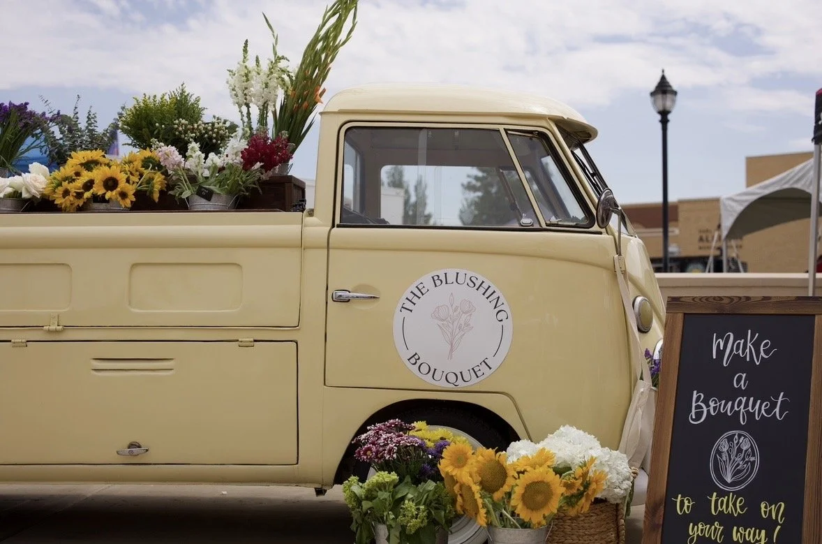 A vintage yellow truck with a flower bouquet and a sign that says "Make a bouquet to take on your way!" at an outdoor market.