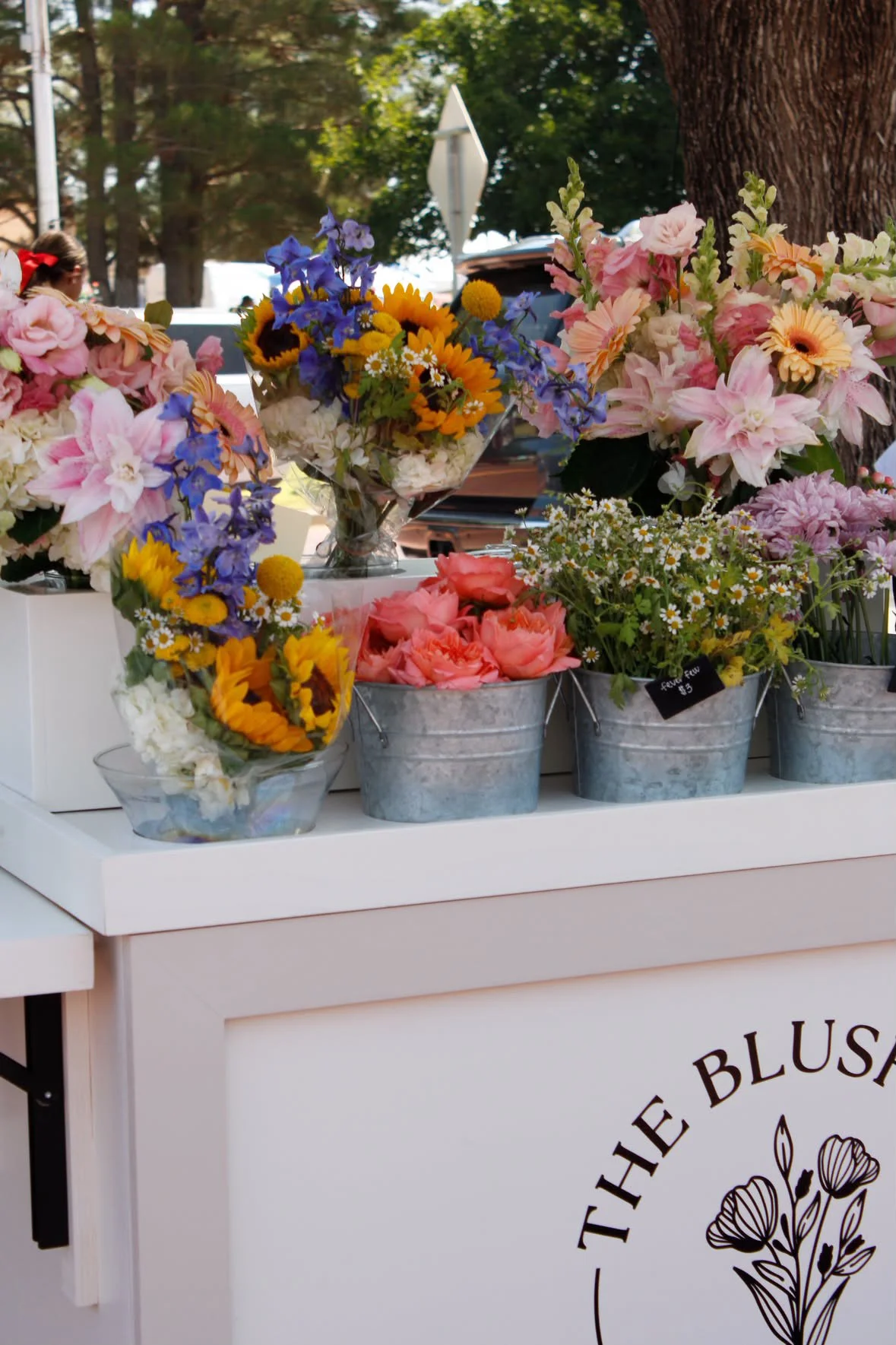 A display of colorful flower bouquets at an outdoor stand, with pink, yellow, purple, and white flowers in metal buckets and a white sign with a floral logo.