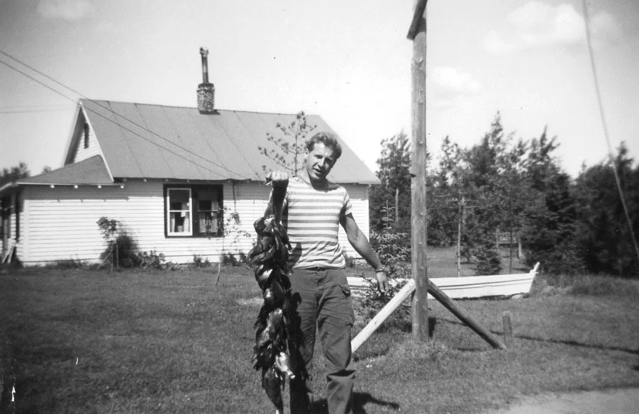 A digital scan of a vintage picture of a man standing outside during daytime, holding a string of dead ducks, with a house, trees, and a boat in the background.