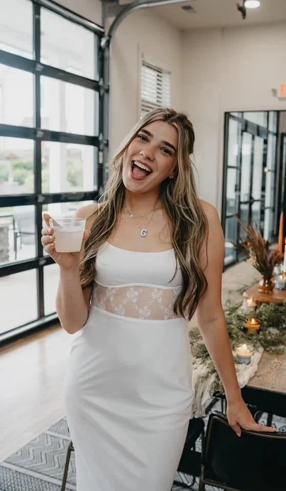 Young woman in a white bridal dress holding a drink and smiling in a modern indoor space with large windows and decorated table.