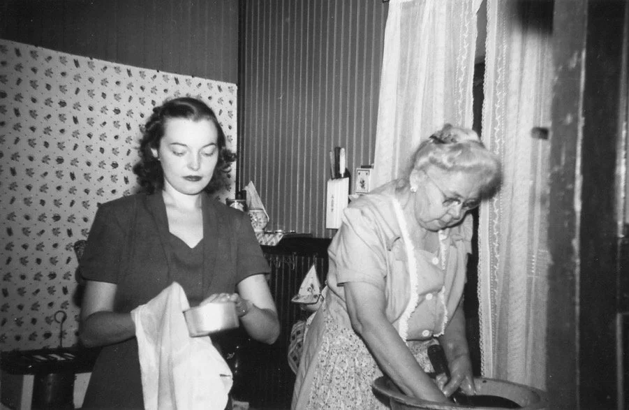 A digital scan of a vintage photo of two women in vintage clothing, one young and one elderly, standing in a room with curtains and patterned wallpaper, engaged in laundry activities.