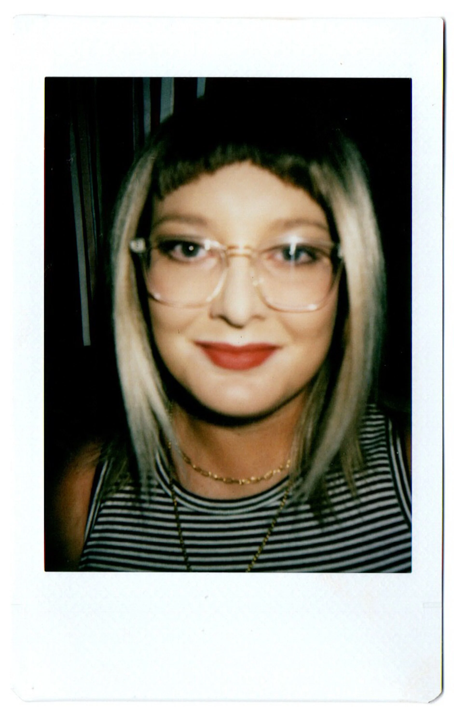 A close-up photograph of Suzy, woman wearing glasses, with blonde hair marked by dark roots and a short black haircut with straight bangs. She is smiling, wearing red lipstick, jewelry, and a black and white striped top. The photo has a white border.