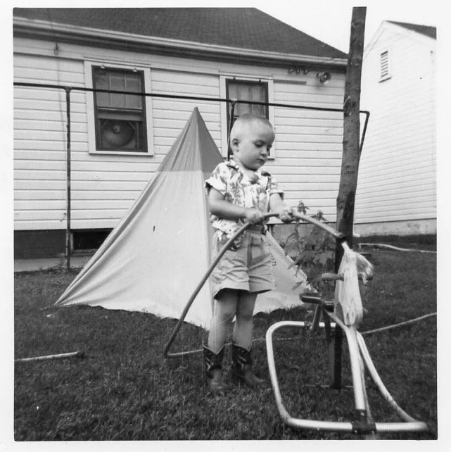 A digital scan of a vintage picture of a young boy standing on grass in front of a small tent in a backyard, holding a stick near a small fountain or water feature, with a house in the background.