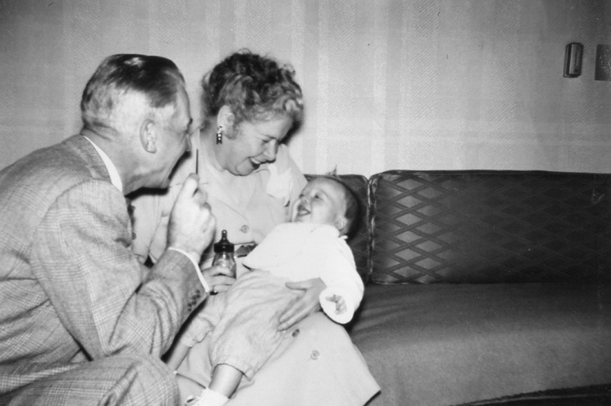 A digital scan of a vintage picture of happy grandparents on a sofa, with a man and woman looking fondly at a smiling baby.