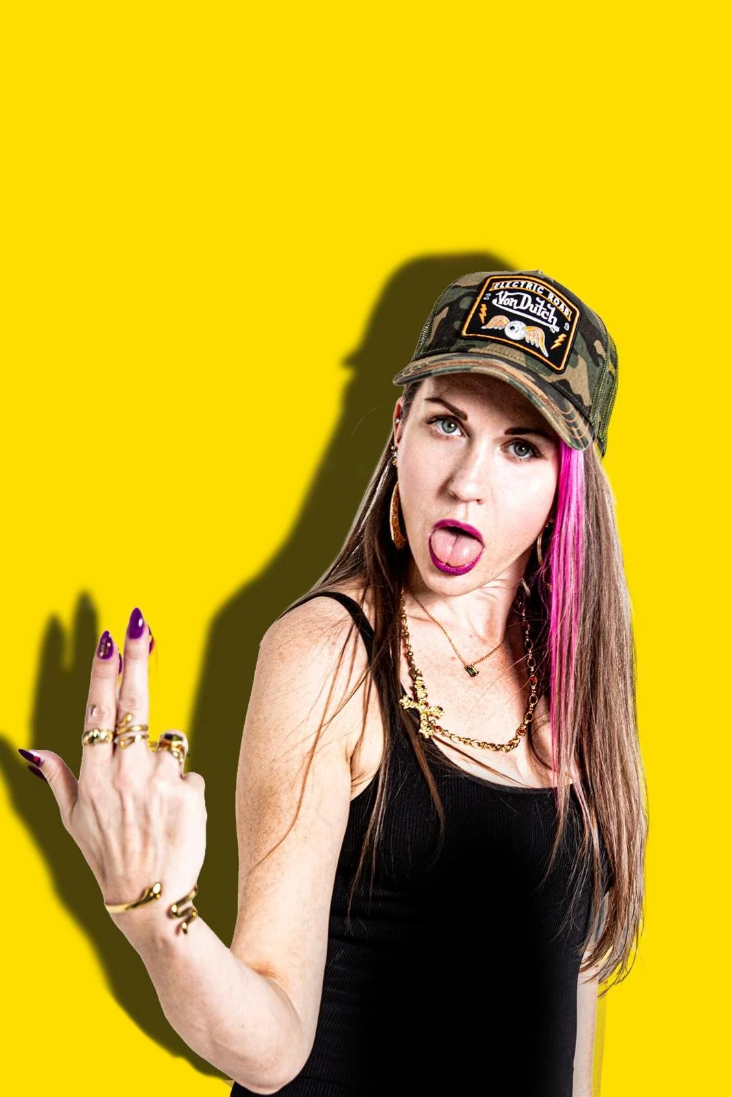 A woman with pink streaks in her hair, wearing a camo cap with patches, black tank top, and gold jewelry, gesturing with her hand against a yellow background.