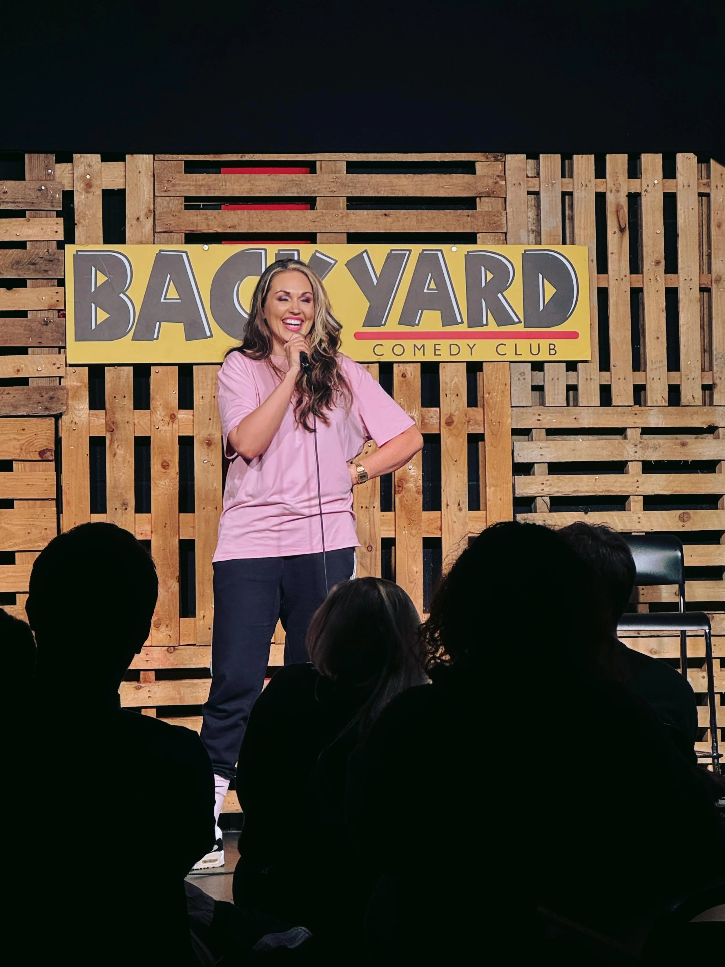 A woman in a pink shirt on stage at the Back Yard Comedy Club, holding a microphone and smiling, with an audience in front of her. The background features a wooden pallet wall and a large yellow sign with the club's name.