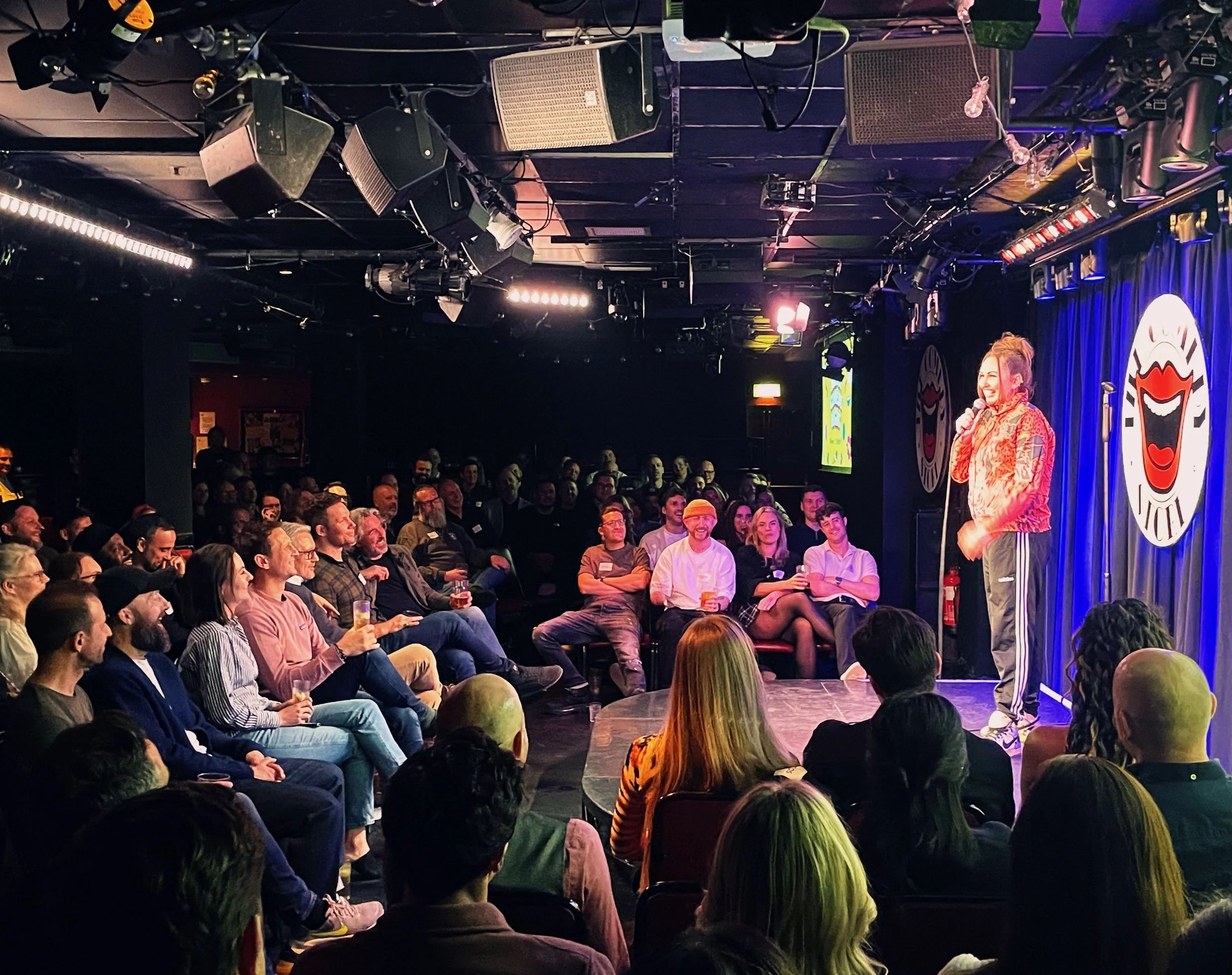 A stand-up comedian performing on stage in front of a large audience at a comedy club. The crowd is engaged and smiling, with some holding drinks. The stage has a curtain with circular artwork and colorful lighting.