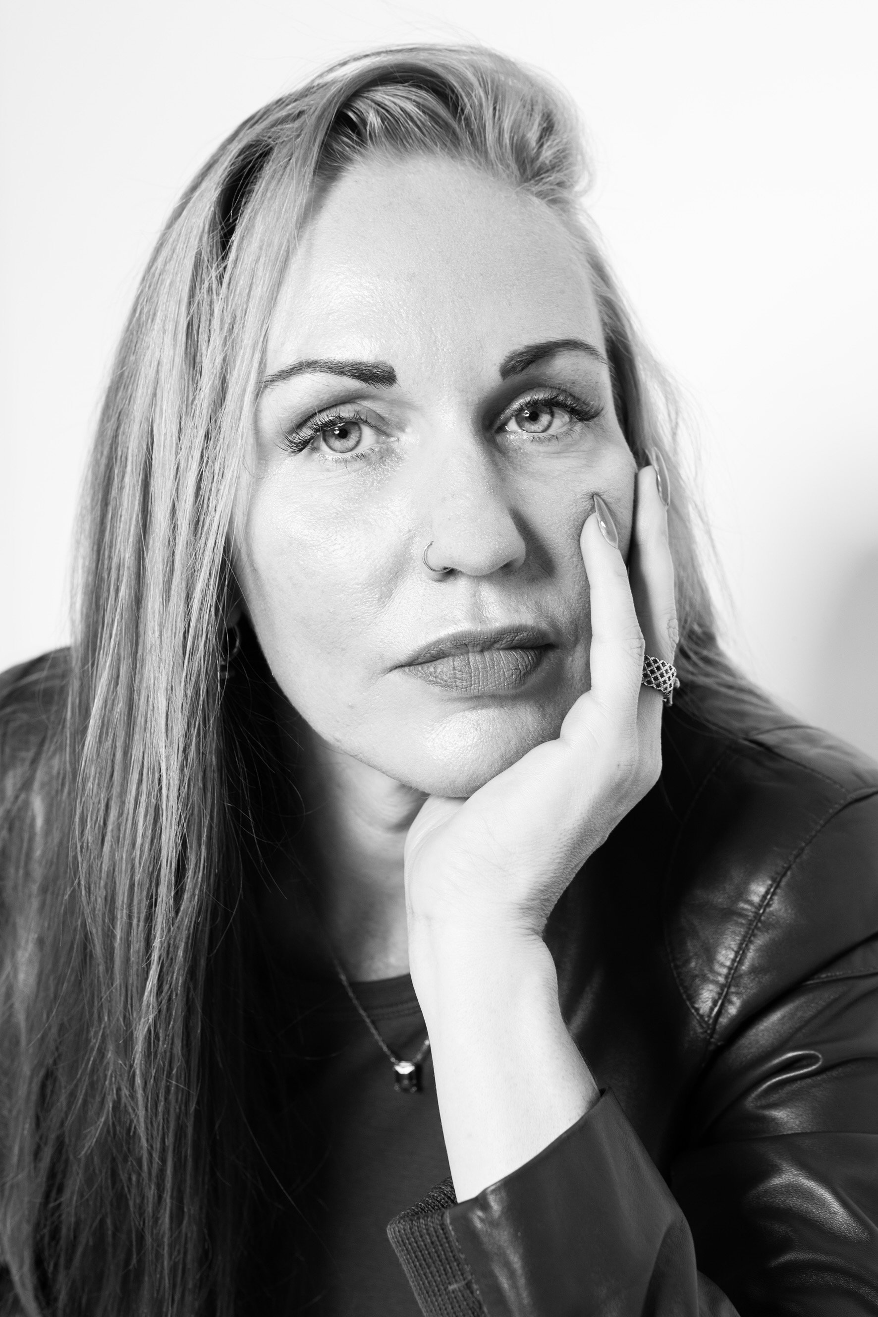 Black and white portrait of a woman with long hair, wearing a ring, a ring on her nose, and a leather jacket, with her hand resting on her face.