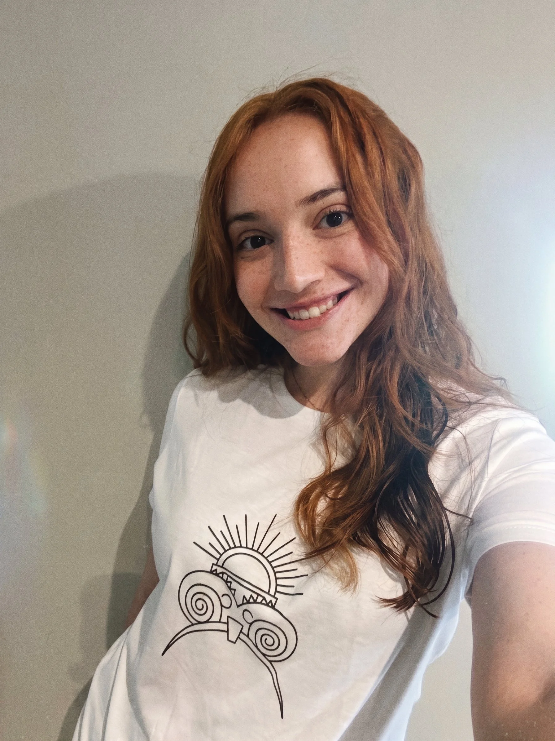 A young woman with long, curly red hair smiling and taking a selfie against a plain wall, wearing a white T-shirt with a black abstract sun or spiral design.