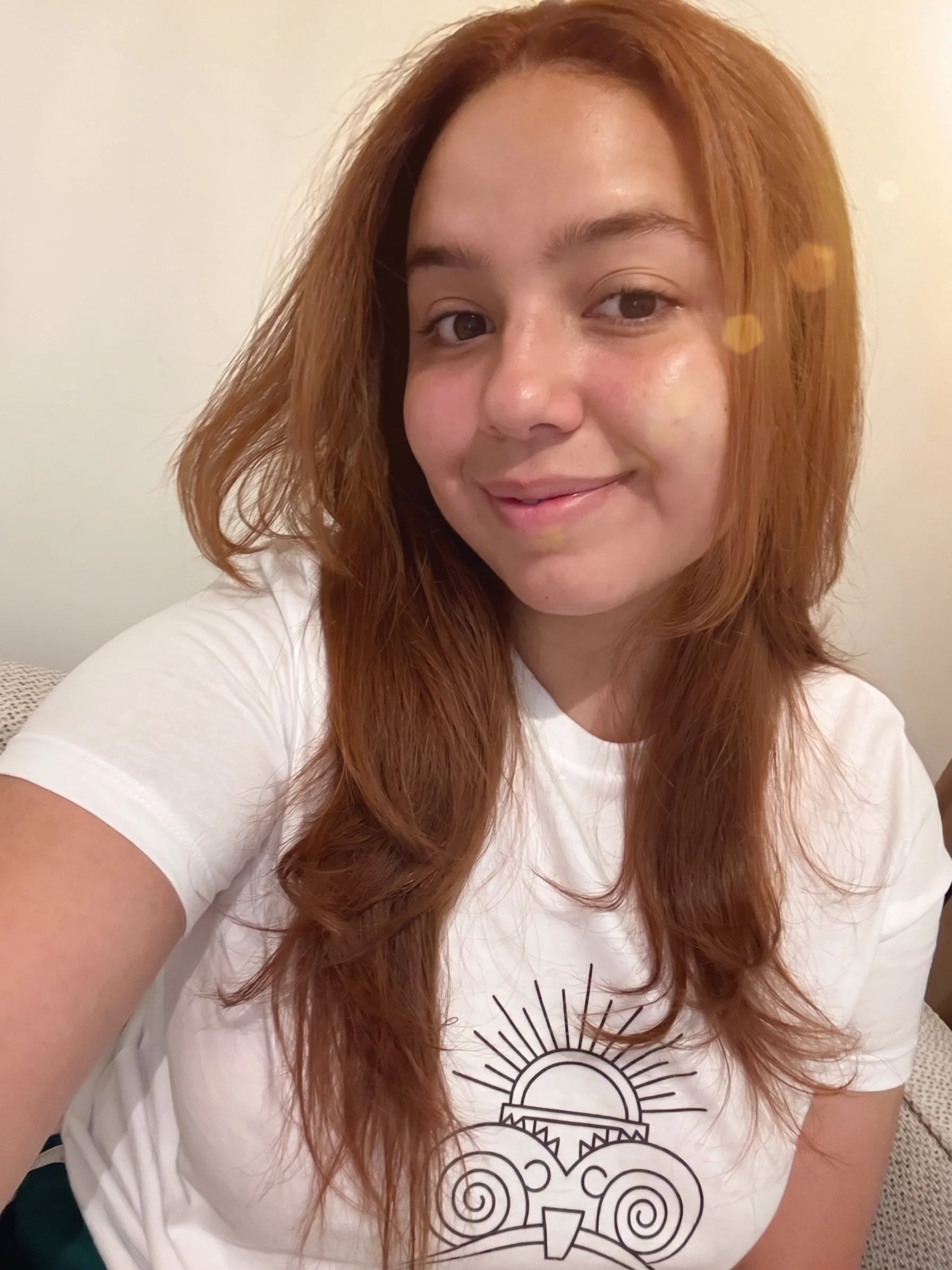A young woman with long, wavy red hair smiling at the camera, wearing a white T-shirt with a graphic design of a stylized sun and intricate geometric patterns.