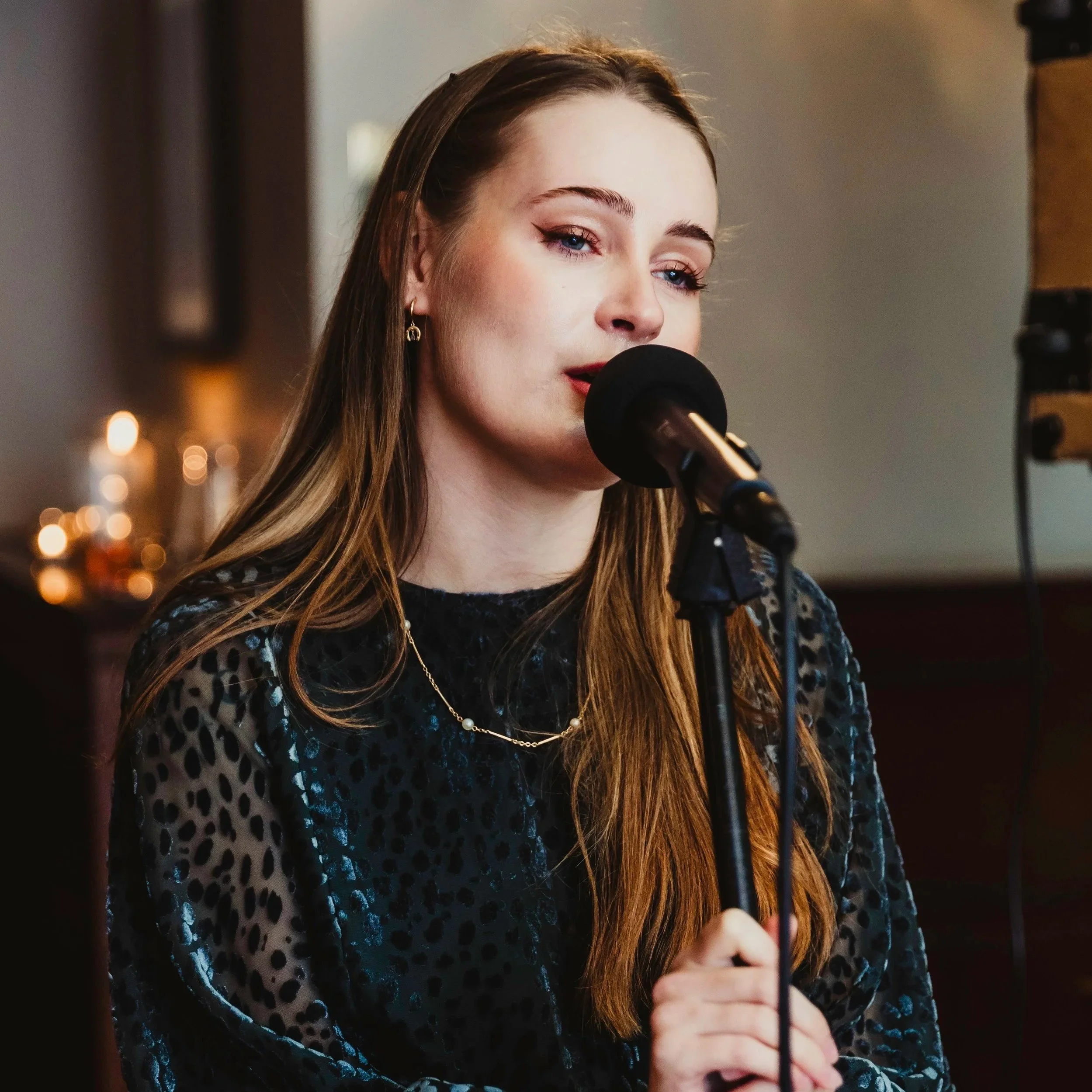 Close-up of a young woman with long brown hair, blue eyes, and makeup, speaking into a microphone at an indoor event, wearing a black and blue patterned dress and a delicate necklace.