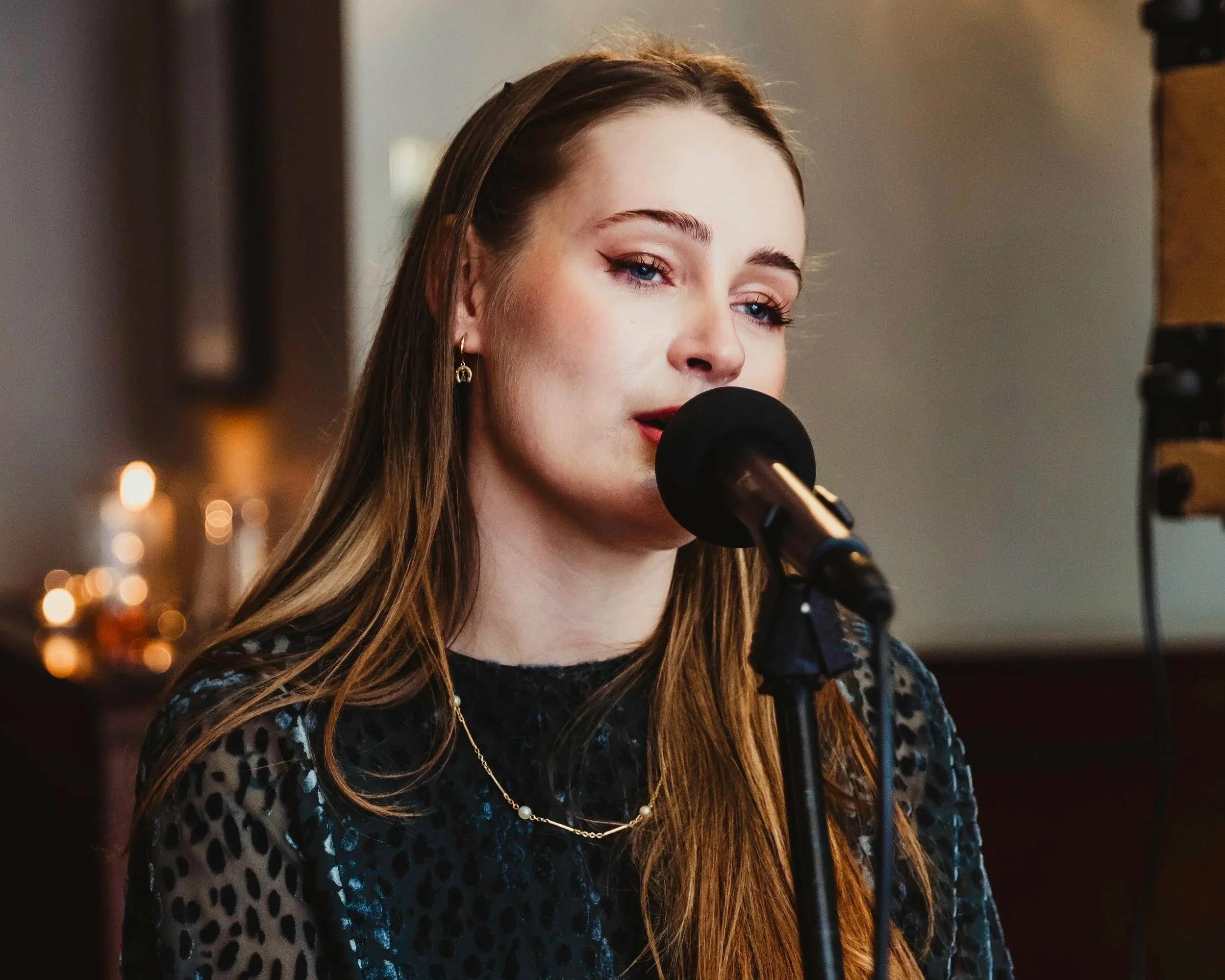Close-up of a woman with long brown hair singing into a microphone, wearing a black and gray patterned top and gold jewelry, with blurred candles in the background.