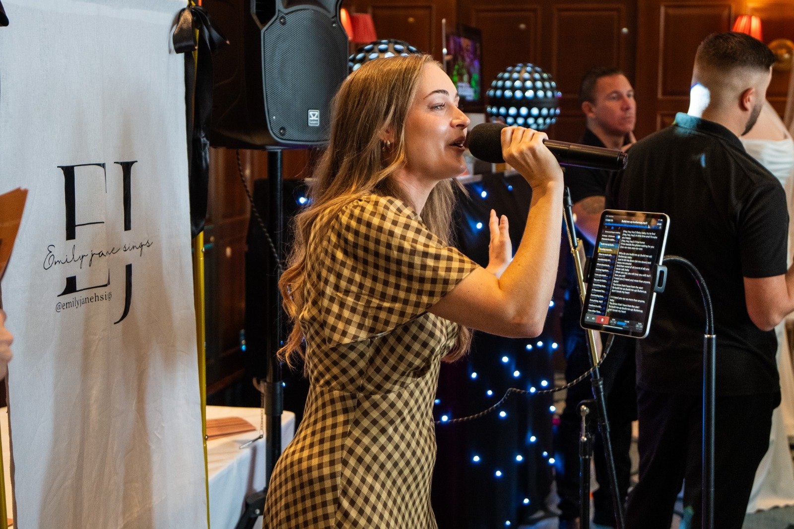 A woman singing into a microphone at an indoor event, with a tablet on a stand displaying song lyrics, and people in the background listening.