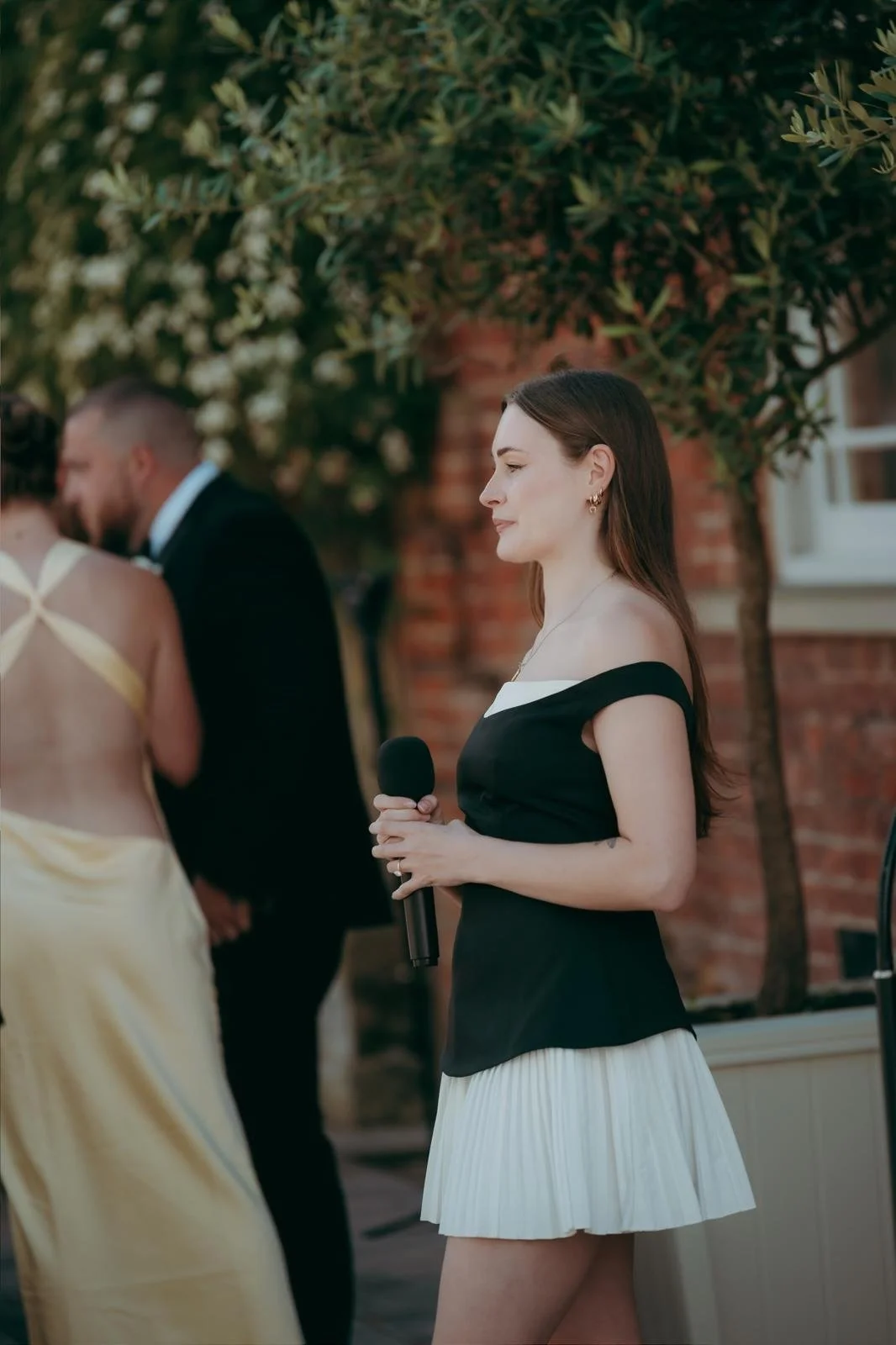 A woman standing outdoors holding a microphone, wearing a black and white off-the-shoulder dress with a pleated white skirt, with people blurred in the background.