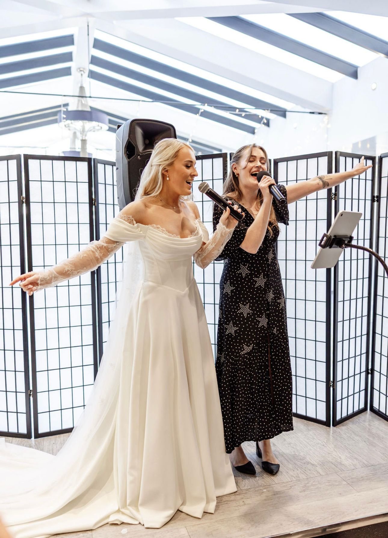 Two women singing into microphones at a celebration or event, with a white and black folding privacy screen behind them and a speaker in the background.