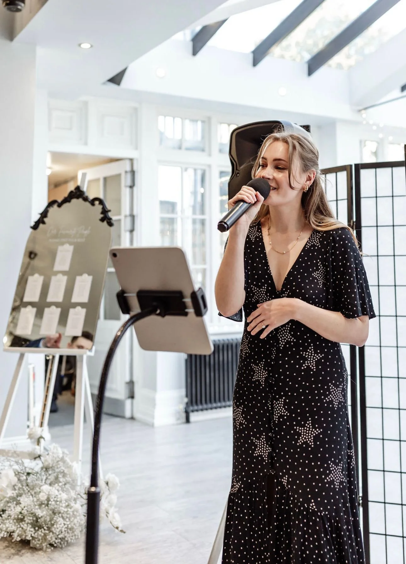A woman in a black dress with white star patterns speaking into a microphone at an event. There is a mirror and a tablet on stands in front of her, and a decorative flower arrangement on the floor, with a room featuring large windows and skylights in the background.