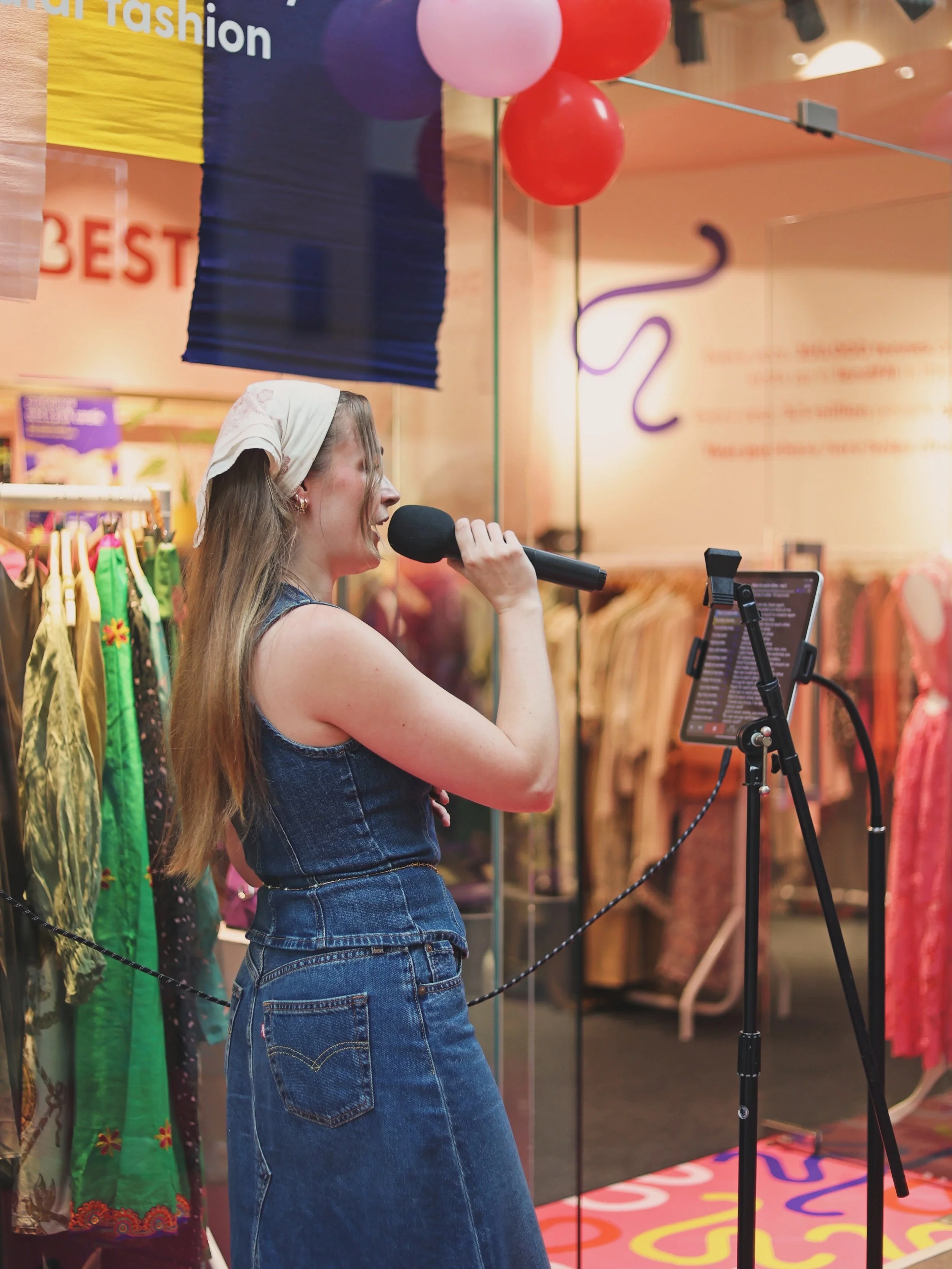 A woman wearing a white headscarf and denim outfit singing into a microphone at an indoor event with colorful balloons, clothing displays, and a sign in the background.