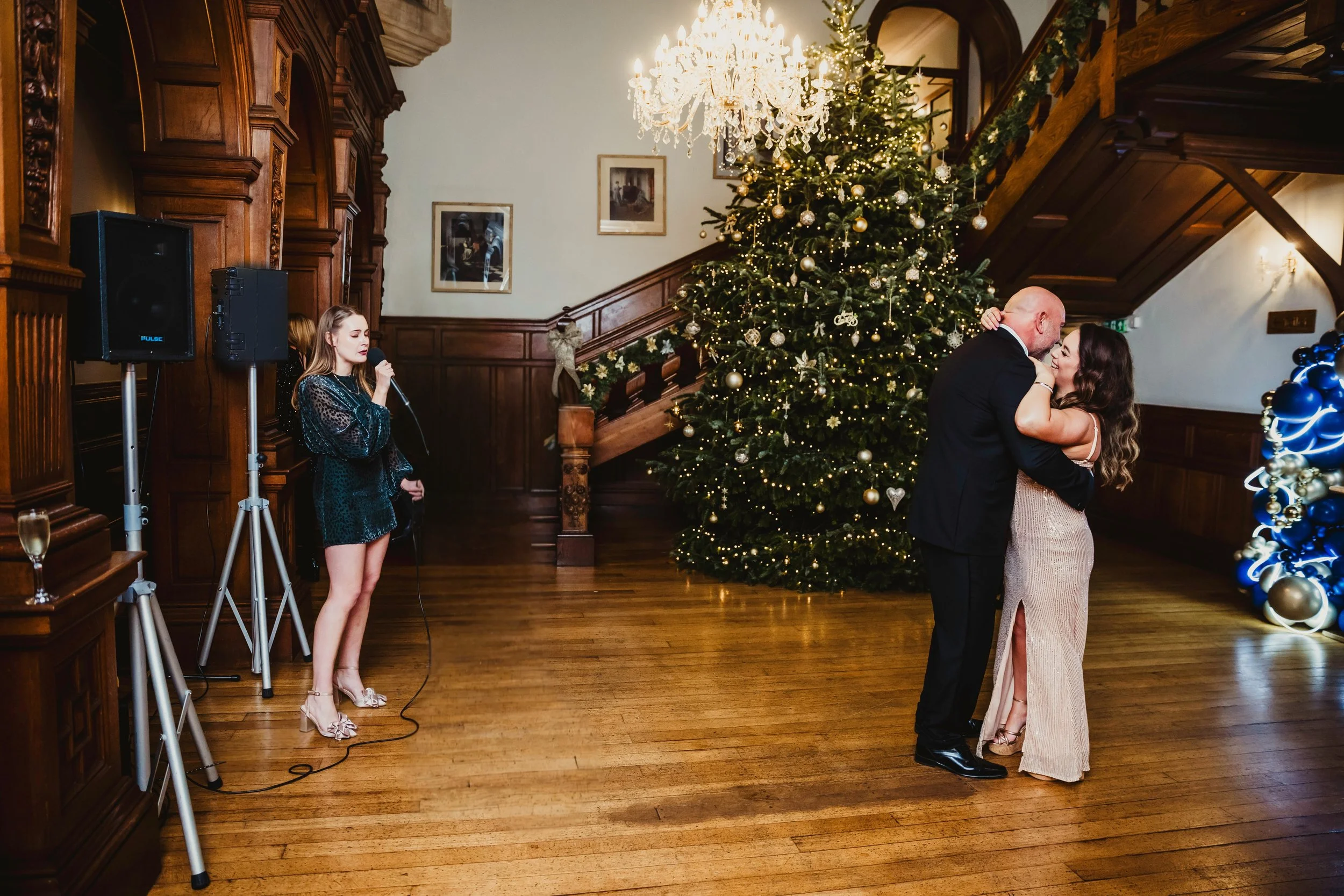 A couple dancing close to each other in front of a decorated Christmas tree in a luxurious, wood-paneled room with a large chandelier. A singer is performing with a microphone nearby.
