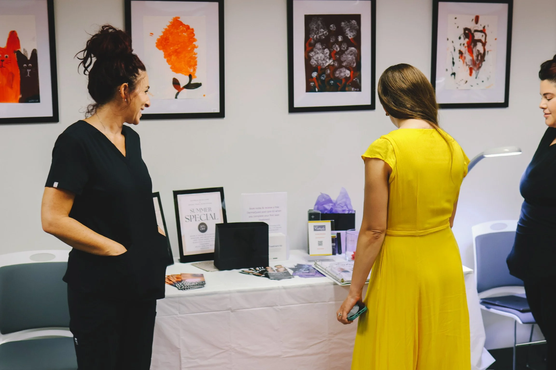 Three women are standing and talking in front of framed artwork displayed on a white wall at an art gallery or event. Two women are facing the artwork, while one woman wearing a black outfit is smiling and engaging with the women. A table with promotional materials and a sign reading 'SUMMER SPECIAL' is visible in the foreground.