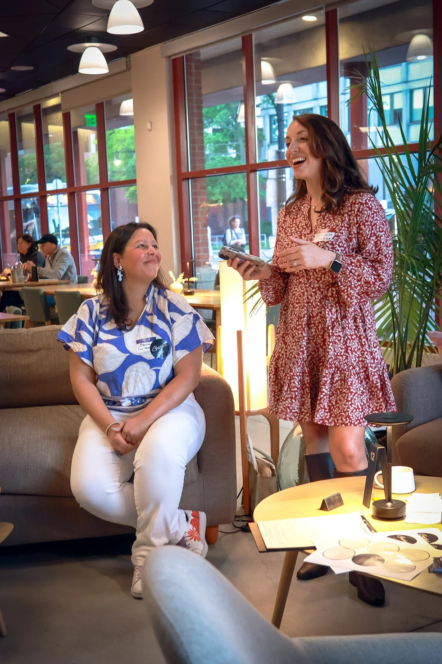 A woman sitting on a sofa looking at another woman who is standing and smiling in a cafe setting. The standing woman is holding a phone and wearing a red floral dress, while the seated woman is wearing a blue and white patterned shirt and white pants.