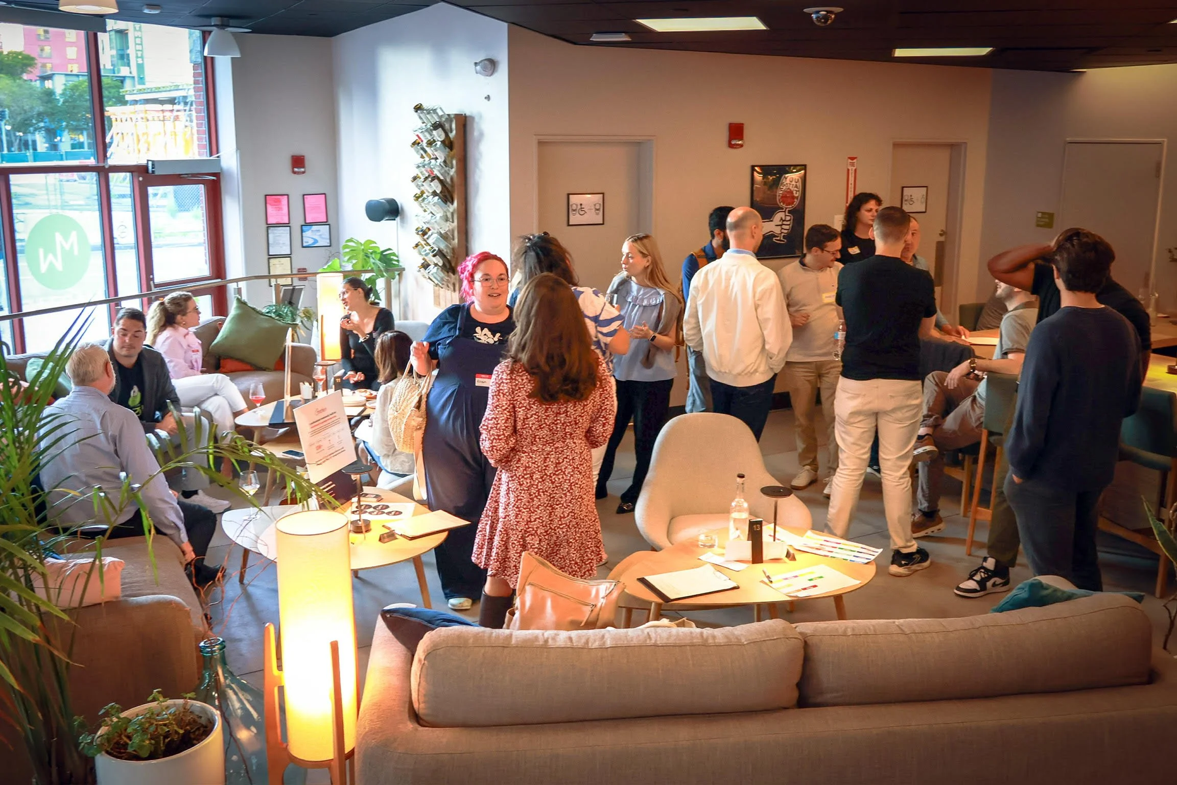 People gathered in a modern indoor space, some standing and talking, others seated at tables, with large windows showing an outdoor scene.