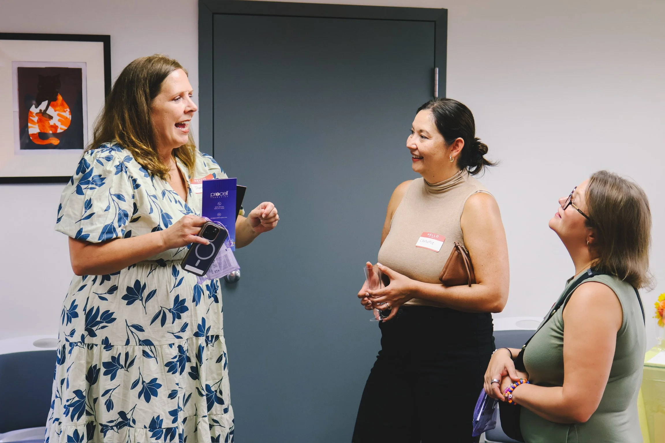 Four women are engaged in conversation at a social event. One woman, wearing a floral dress, is speaking and holding a brochure and a smartphone. The other women are listening, with one holding a glass, and all seem to be smiling and enjoying the interaction.