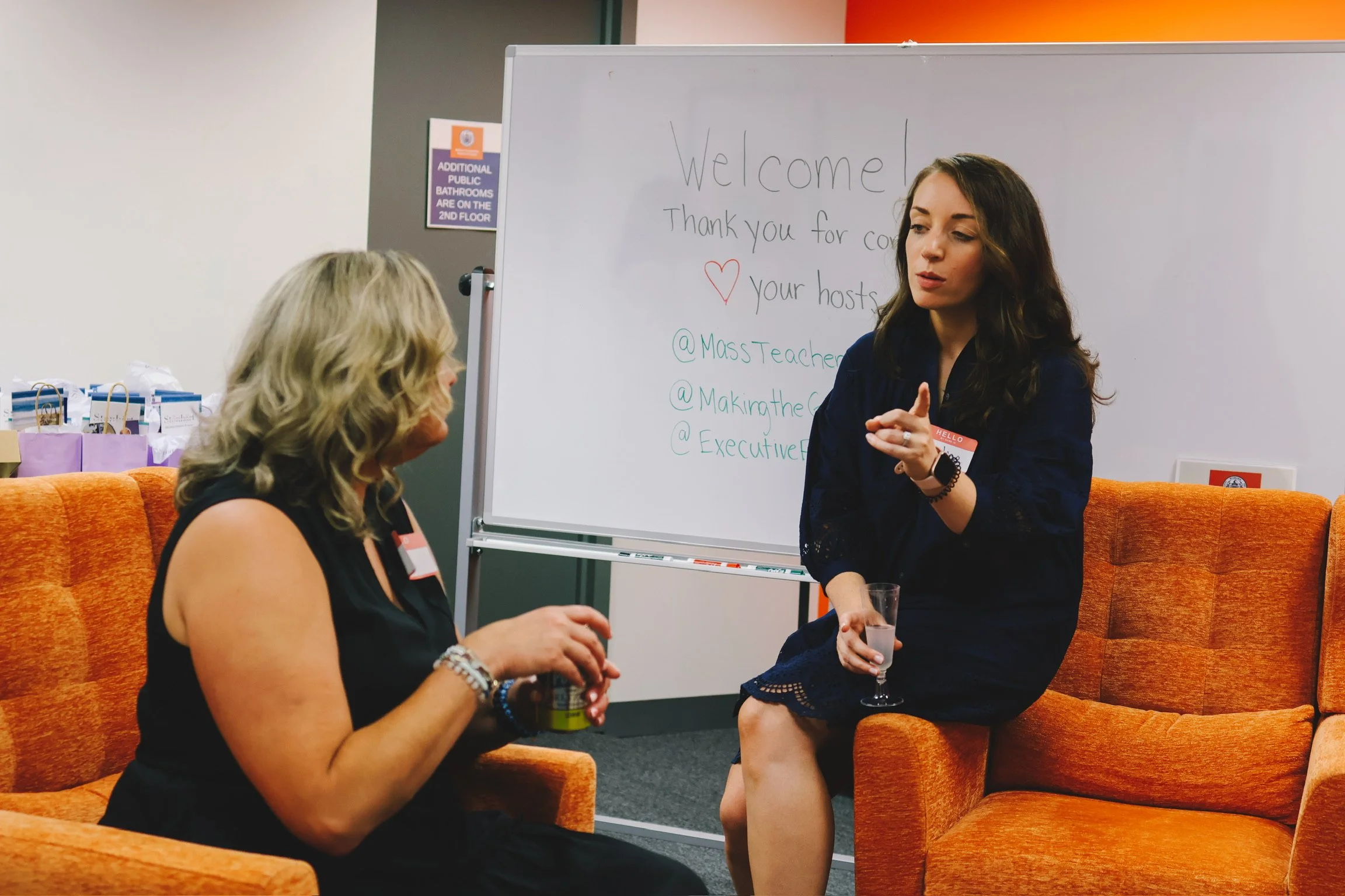 Two women having a conversation at an event, sitting on orange couches in front of a whiteboard with writing on it, holding drinks, with a sign about bathrooms in the background.