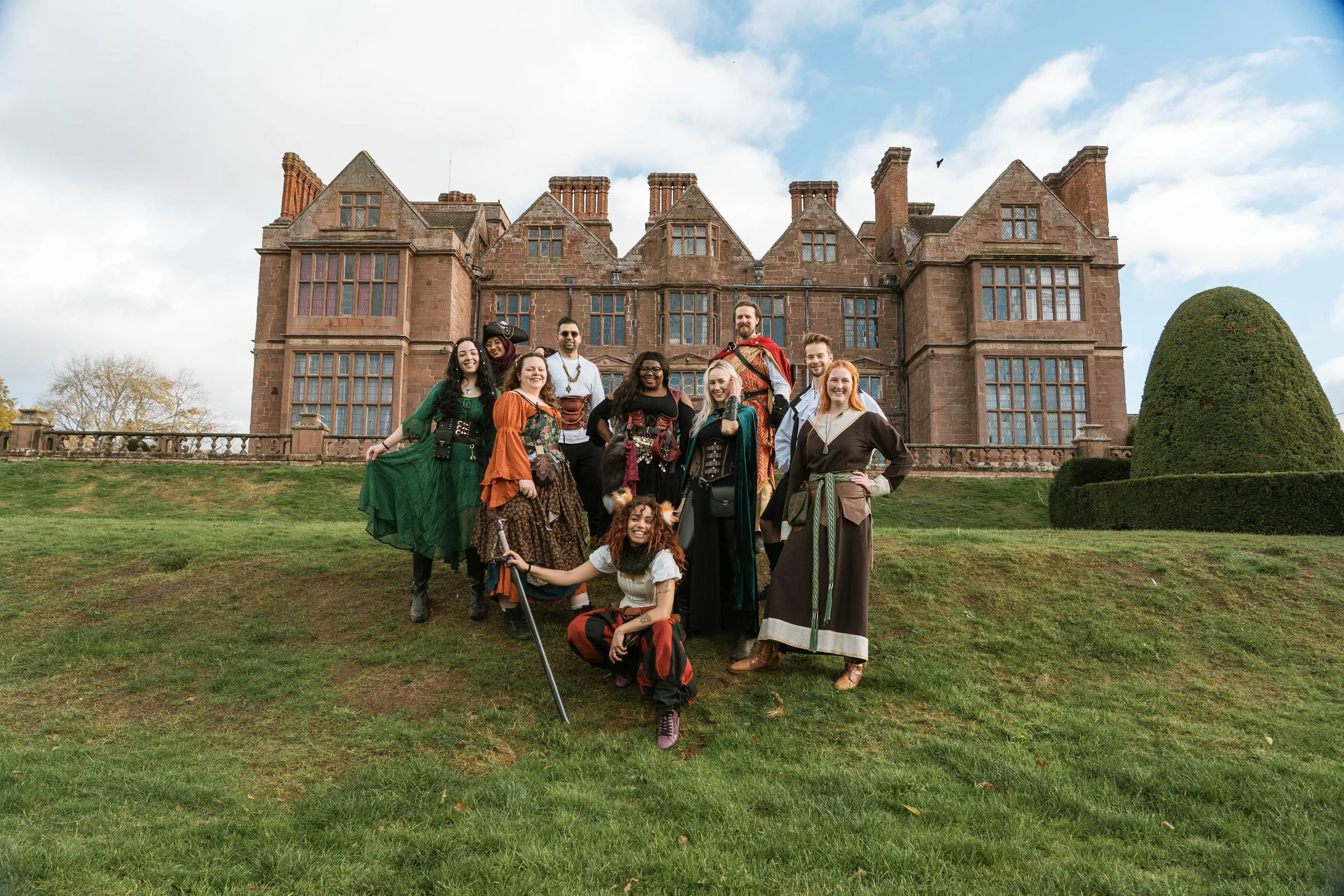 Group of nine people dressed in medieval or fantasy costumes posing in front of a large castle with pointed towers and multiple windows.