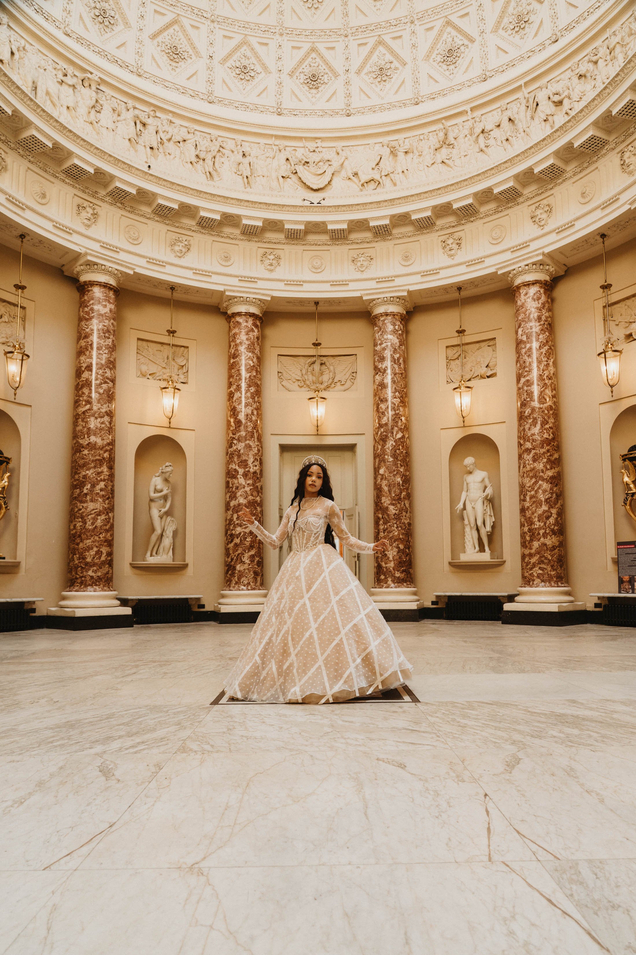 A woman in an elegant beige gown standing in a grand, classical-style hall with marble columns, sculptures, and ornate ceiling reliefs.