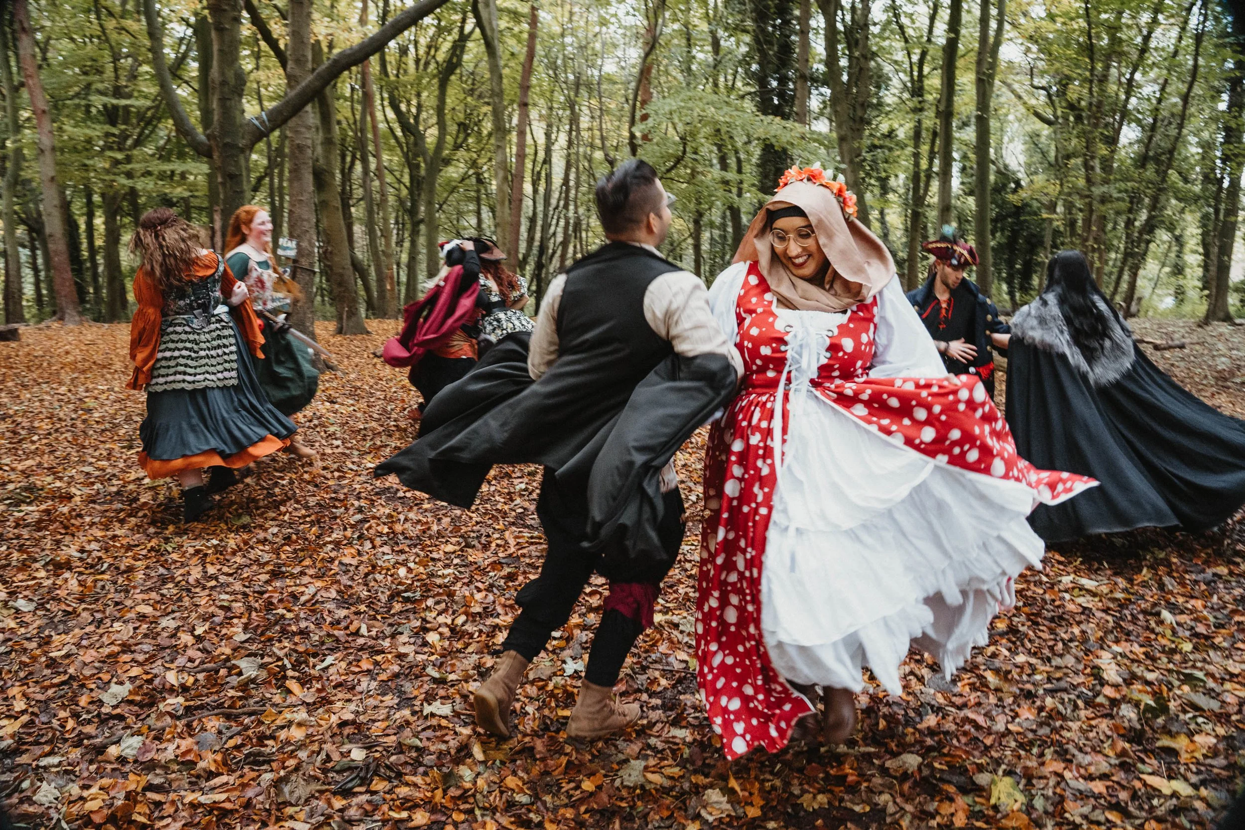 People in historical costumes dancing in a forest with fallen autumn leaves.