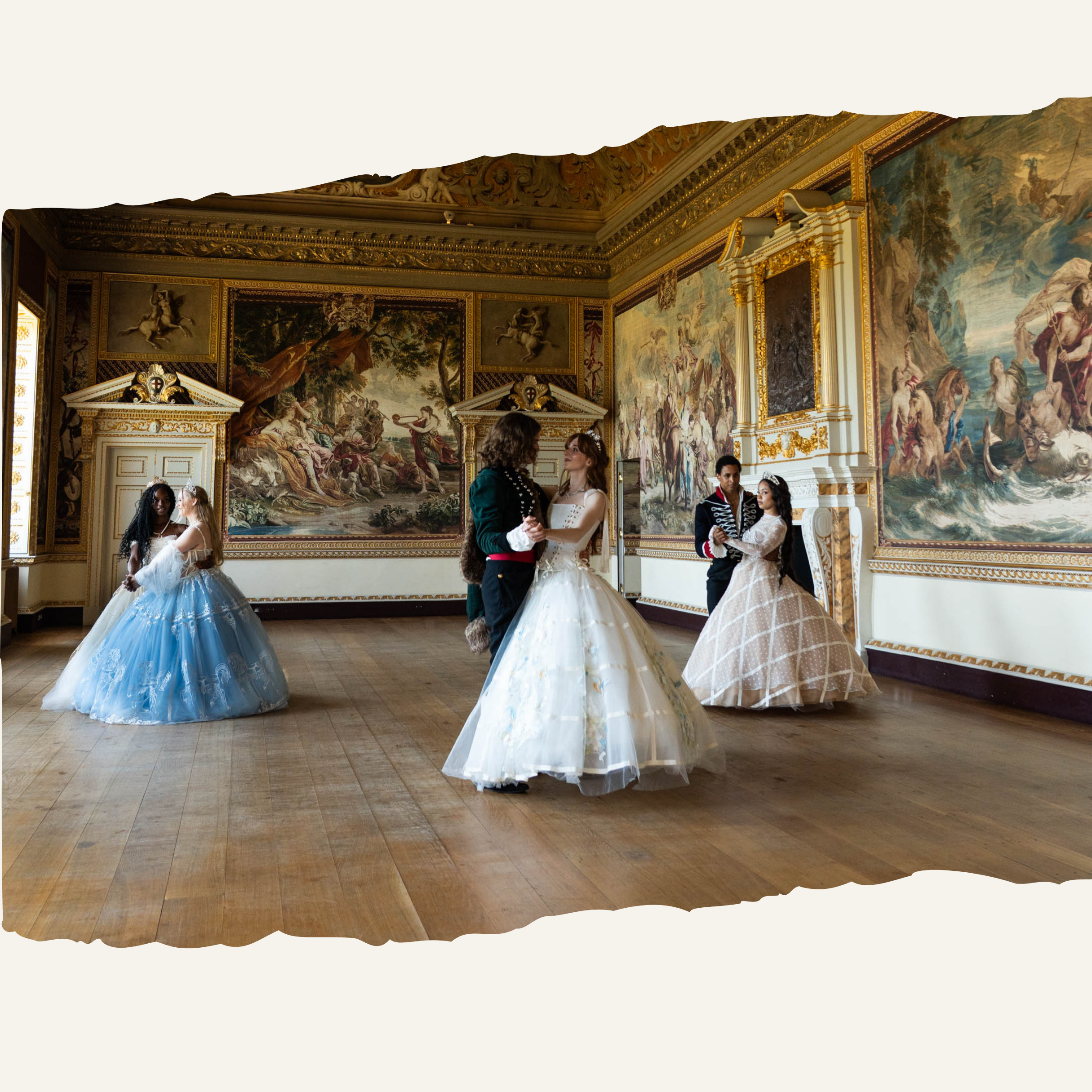 Group of people dressed in historical costumes dancing in a richly decorated room with ornate gold trim and large scenic paintings.