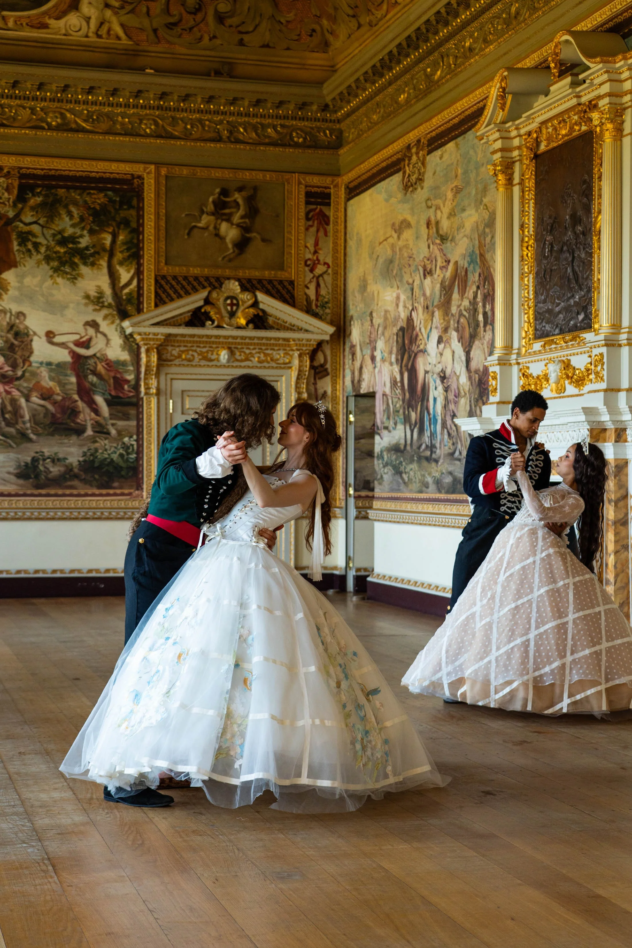 Two couples in vintage formal attire dancing in a grand, ornate room with elaborate tapestries and gold accents.