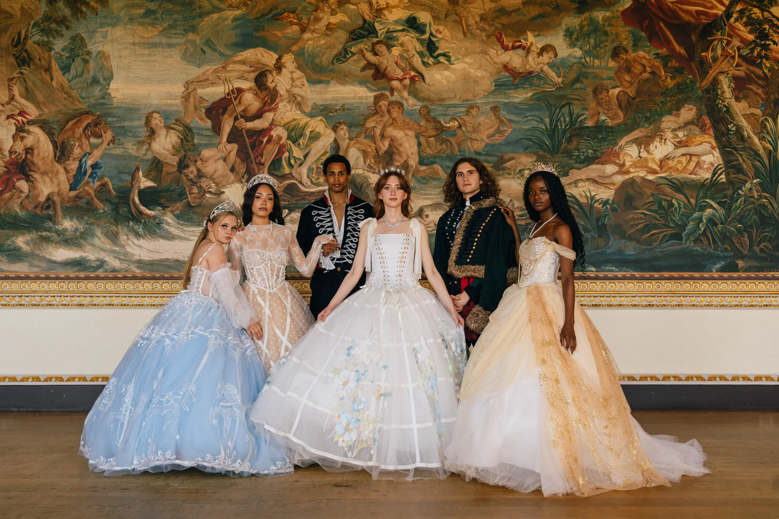 Six women dressed as princesses in princess gowns posing in front of a large classical painting with mythological figures and clouds.