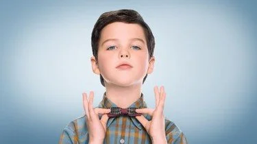 A young boy with dark hair, wearing a checkered shirt, holds a large bow tie in front of his neck.