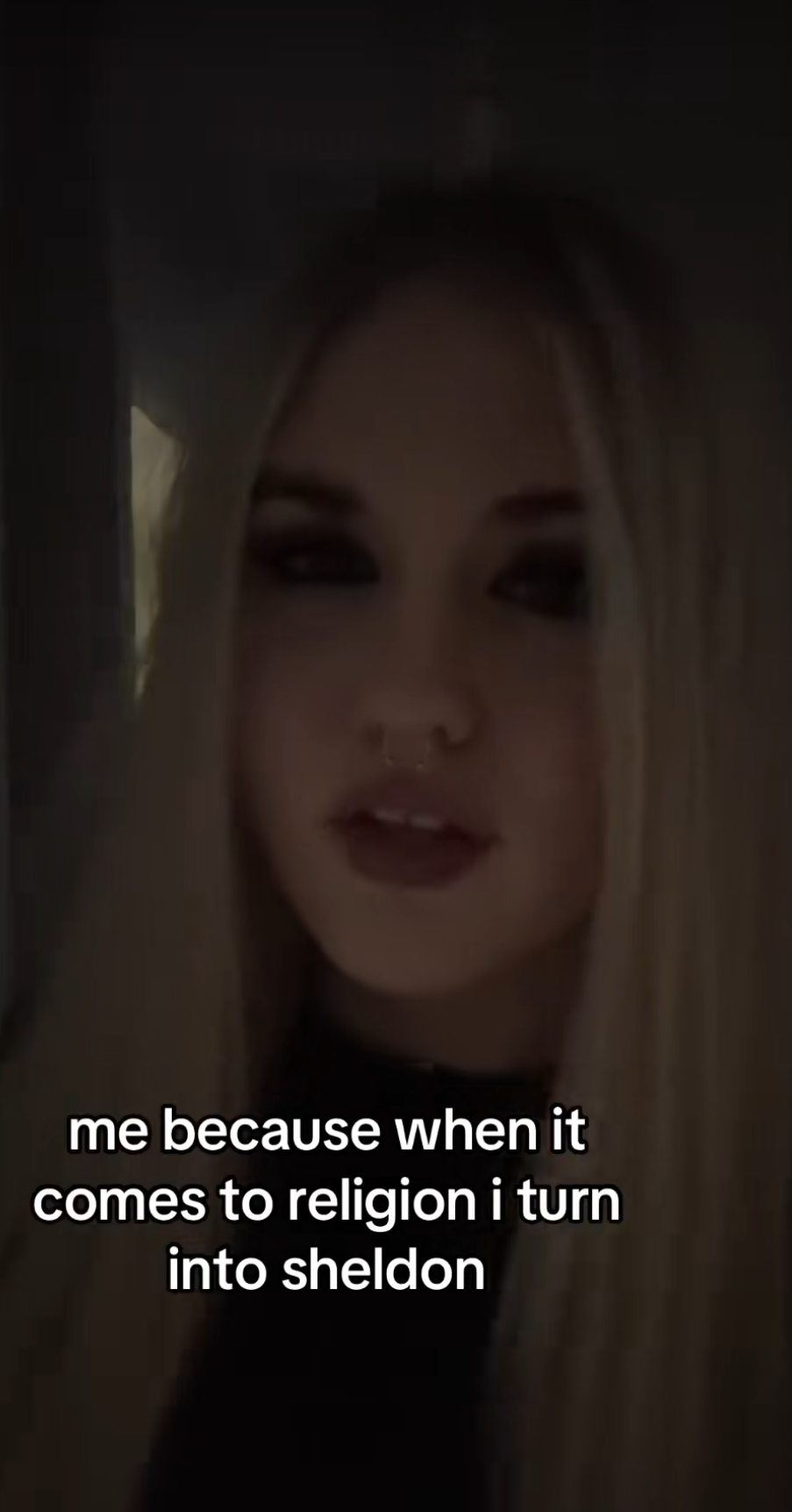 A woman with long blonde hair, dark eye makeup, and a septum piercing, smiling slightly in a dimly lit environment. Text overlay reads: 'me because when it comes to religion I turn into sheldon'.