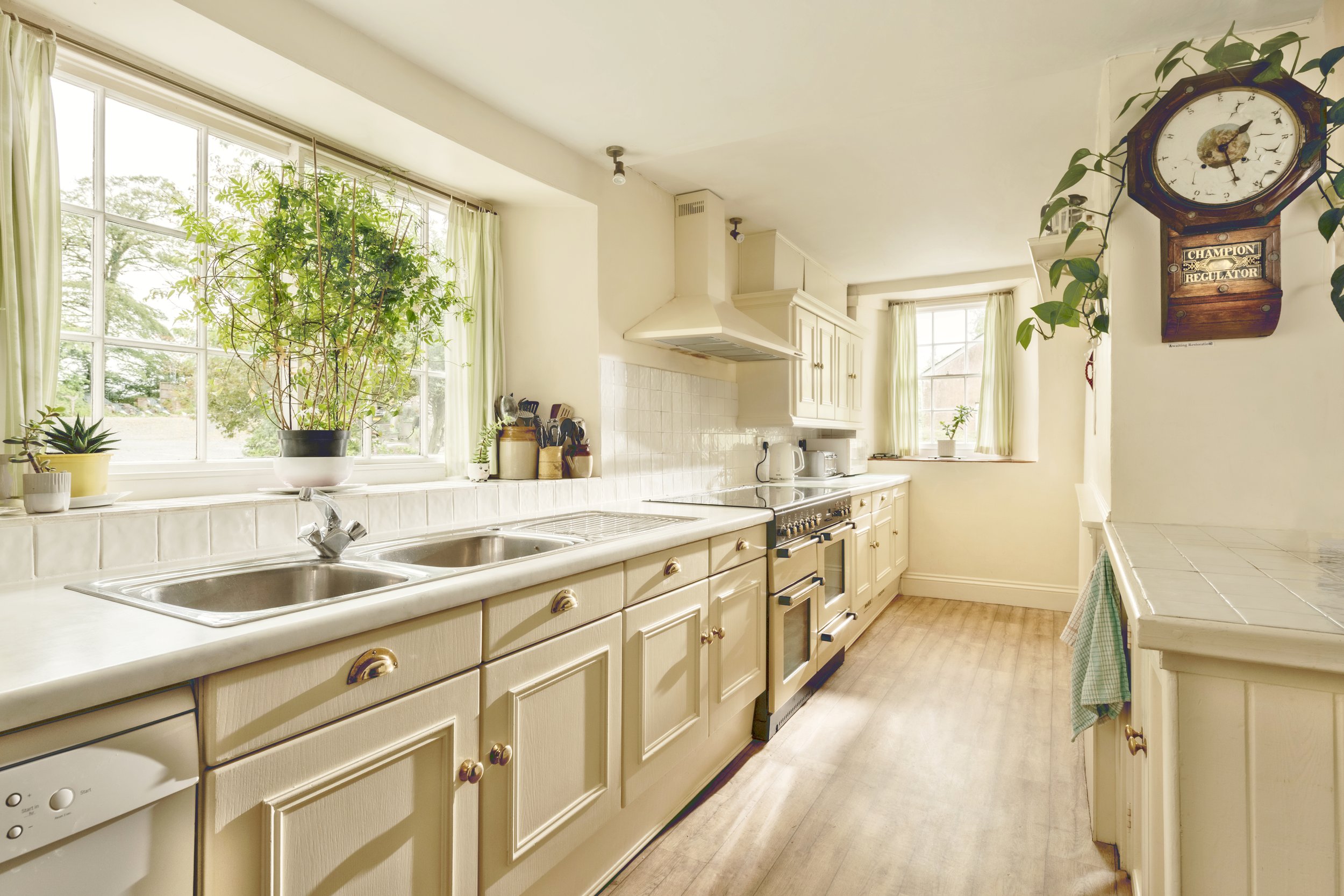 A cozy kitchen with large windows, cream-colored cabinets, and a white tiled countertops. Potted plants sit on the window sill, and a vintage wall clock is mounted on the wall near a leafy vine at Parnacott house.