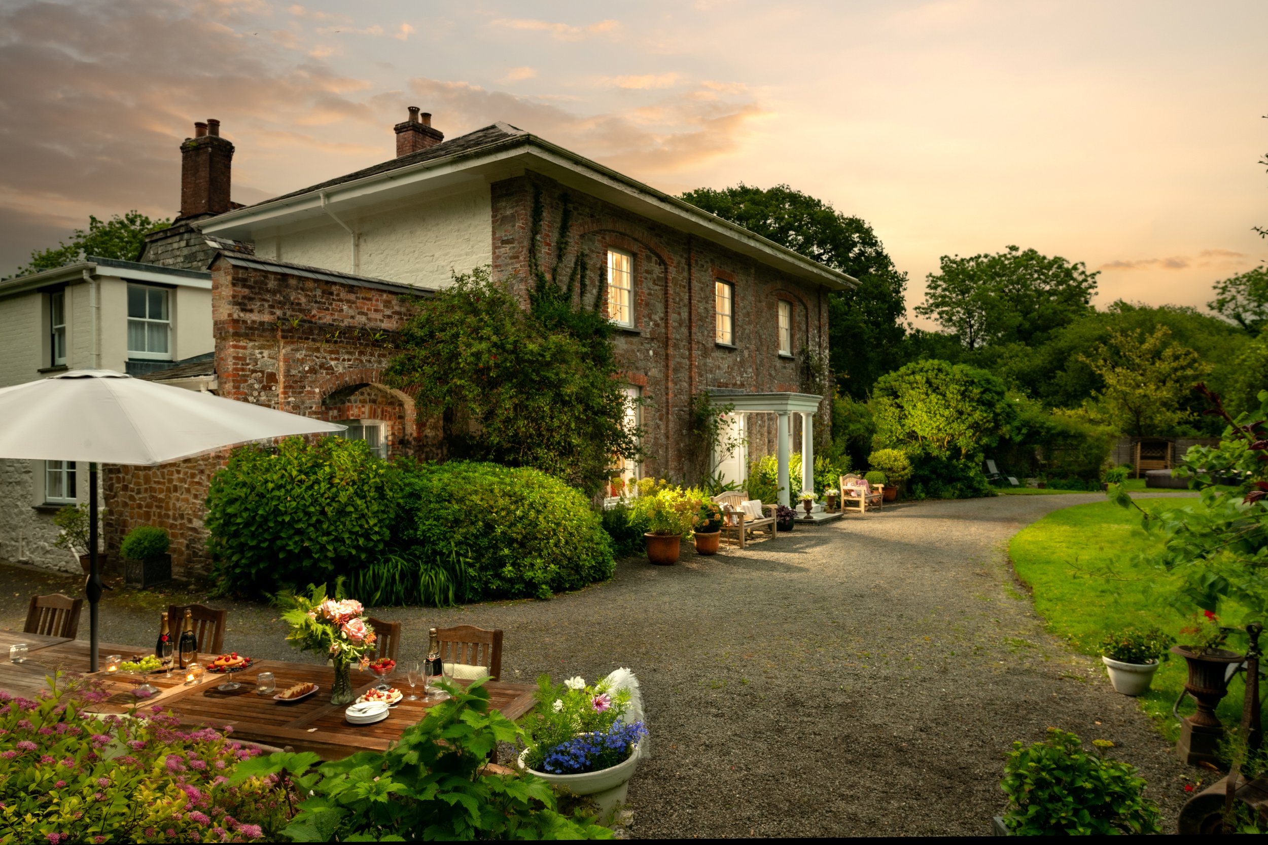 A brick house with multiple chimneys, trees, and a garden with potted plants. There is an outdoor table set with food, flowers, and a large white umbrella, suggesting a gathering or outdoor meal during sunset or early evening at Parnacott house.