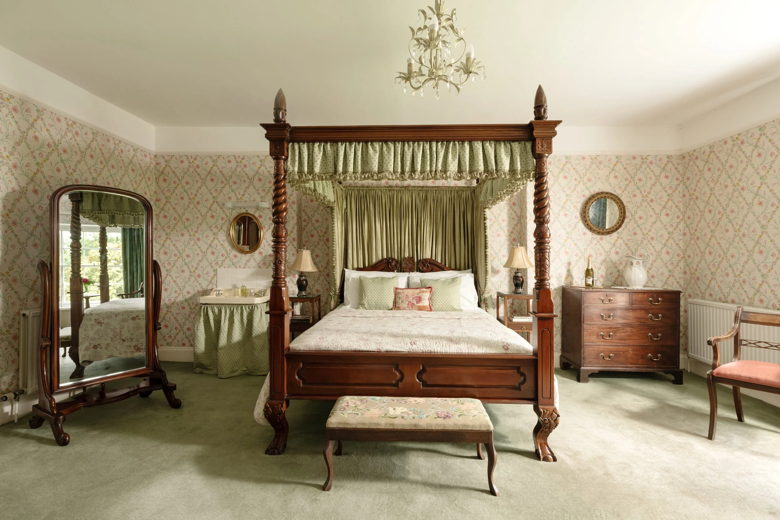 A vintage master bedroom with a four-poster wooden bed, floral wallpaper, green curtains, a large mirror, and antique furniture at Parnacott house. 