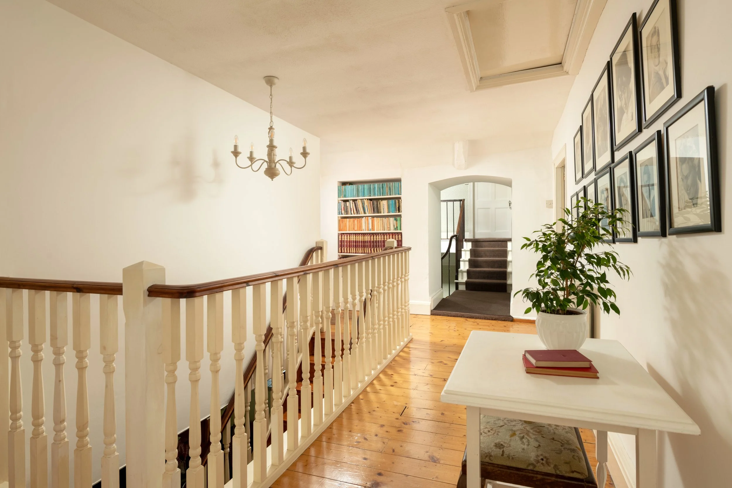 Interior view of a home staircase landing with a chandelier, bookcase, framed pictures, and a table with books and a potted plant at Parnacott house.