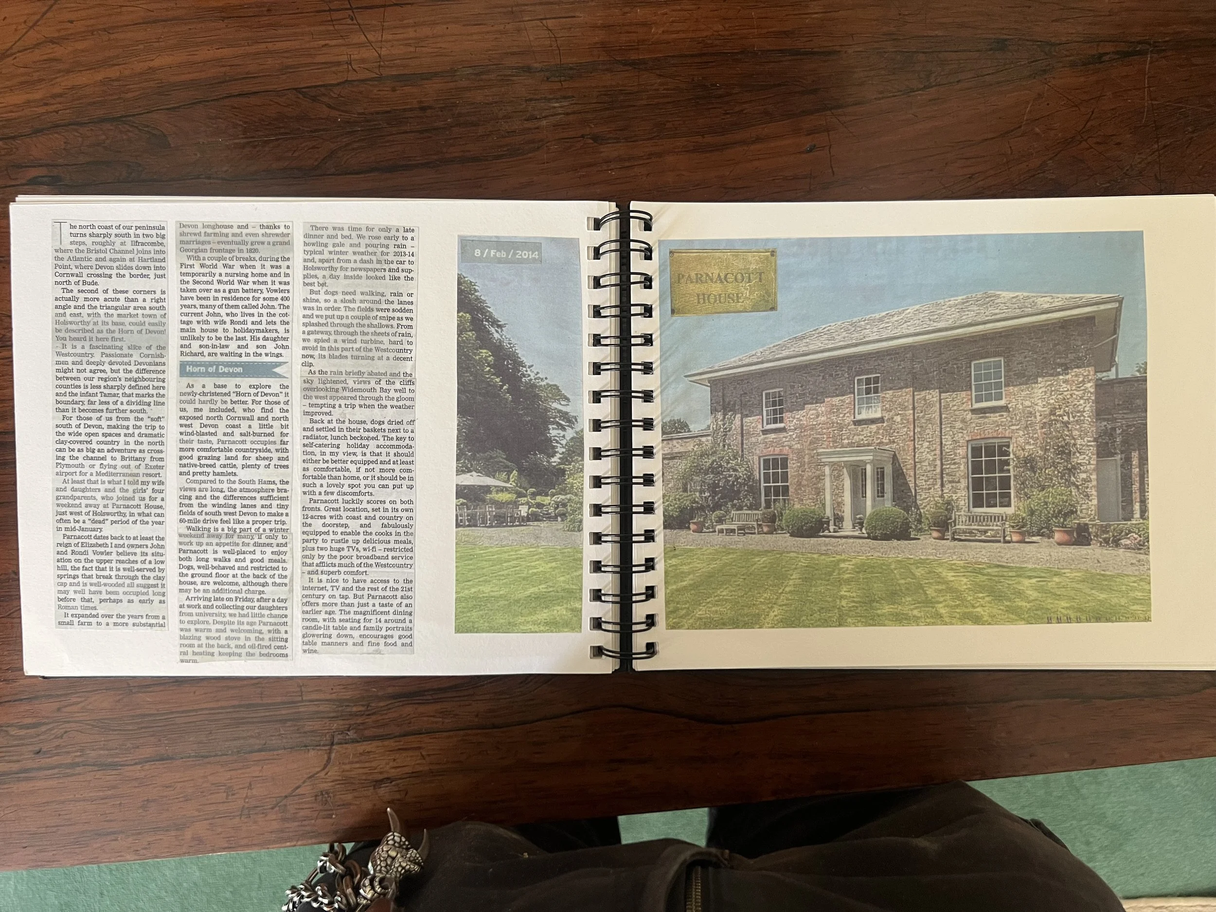 Open spiral-bound book displaying two pages; the left page contains text from a travel or history article, and the right page shows a color photograph of a stone house with multiple windows and a well-maintained lawn with benches and potted plants. The top of the right page has a small section with the date '8 / Feb / 2014' and a label reading 'PARNACOTT HOUSE.' The book is placed on a wooden table, and the person taking the photo is wearing black pants and sneakers.